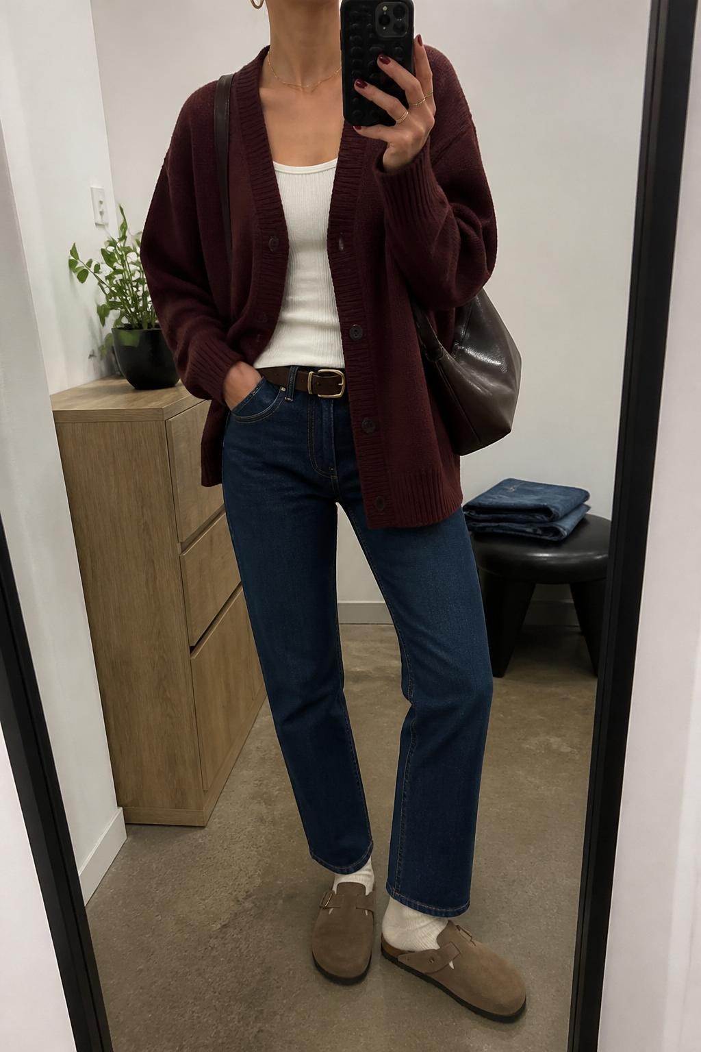 mirror selfie of a woman wearing the exact outfit described above, clearly showing mocha suede Birkenstock-style clogs with ivory socks, dark blue straight-leg jeans, a fitted ivory tank, a burgundy oversized button-front cardigan, a chocolate brown shoulder bag, and gold huggie earrings, holding a phone in front of a mirror, cropped at NECK so the face does not show, with a relaxed casual selfie pose, in a minimal dressing room, soft indoor lighting, with a small plant on a dresser and folded denim jeans on a bench, latest iPhone photo quality.