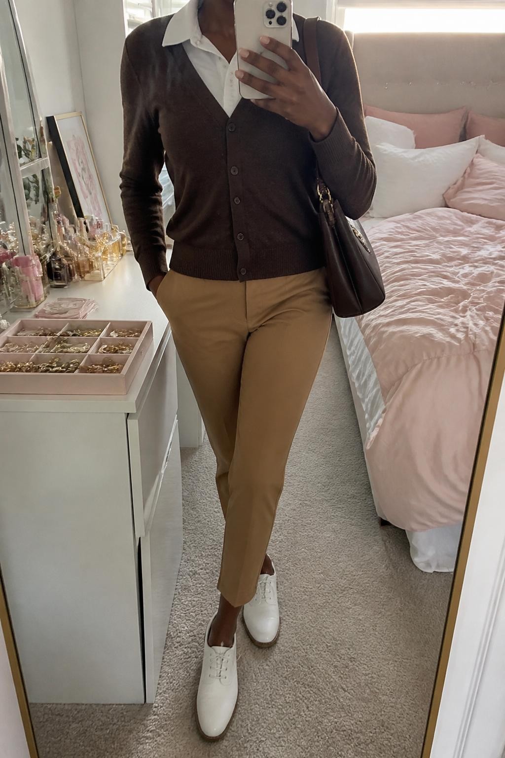 Mirror selfie of a Black woman wearing a fitted chocolate brown button-front golf cardigan over a white short-sleeve collared polo, paired with camel ankle-length golf trousers, clearly showing white leather golf shoes, a tan visor, and a structured chocolate brown shoulder bag, holding an iPhone in front of a feminine-themed bedroom mirror, cropped at the neck so the face does not show, relaxed casual selfie pose, natural window light, a jewelry tray on the vanity and soft pillows on an unmade bed, natural unstaged setting, latest iPhone photo quality.