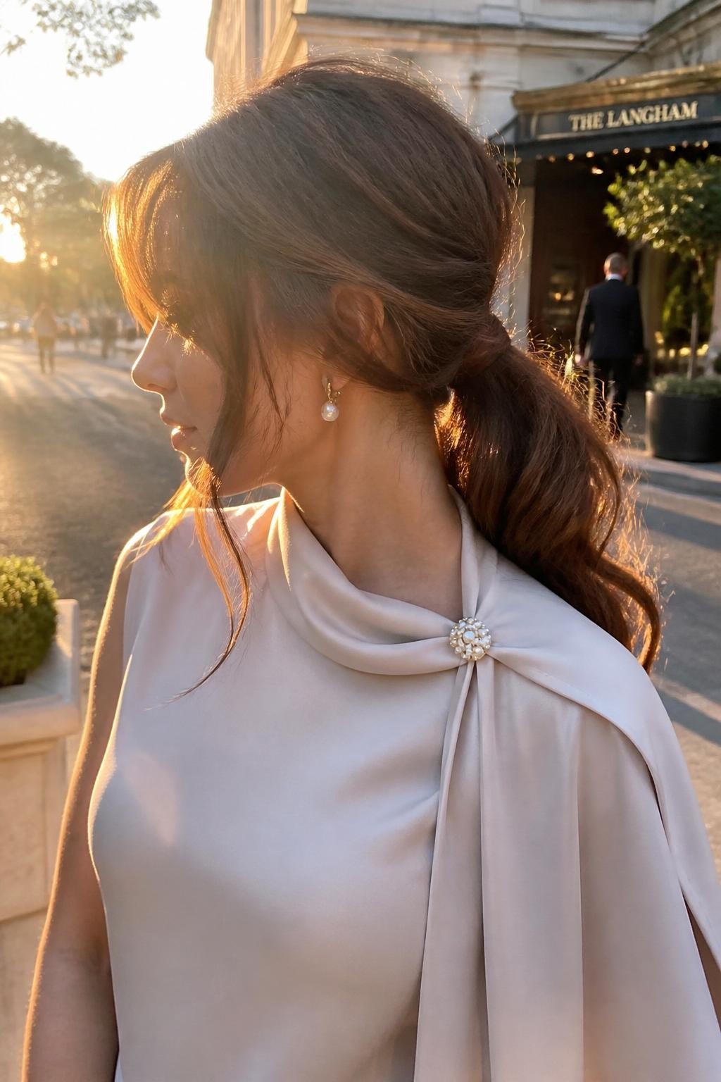 Image prompt: Photo of a woman wearing a loose low ponytail with curled lengths, soft crown volume, and two face-framing curls, side profile view, softly blurred city street outside a wedding hotel, golden hour glow, containing pearl earrings and a small hair clip attached to her dress collar, realistic candid wedding guest moment, latest iPhone photo quality.