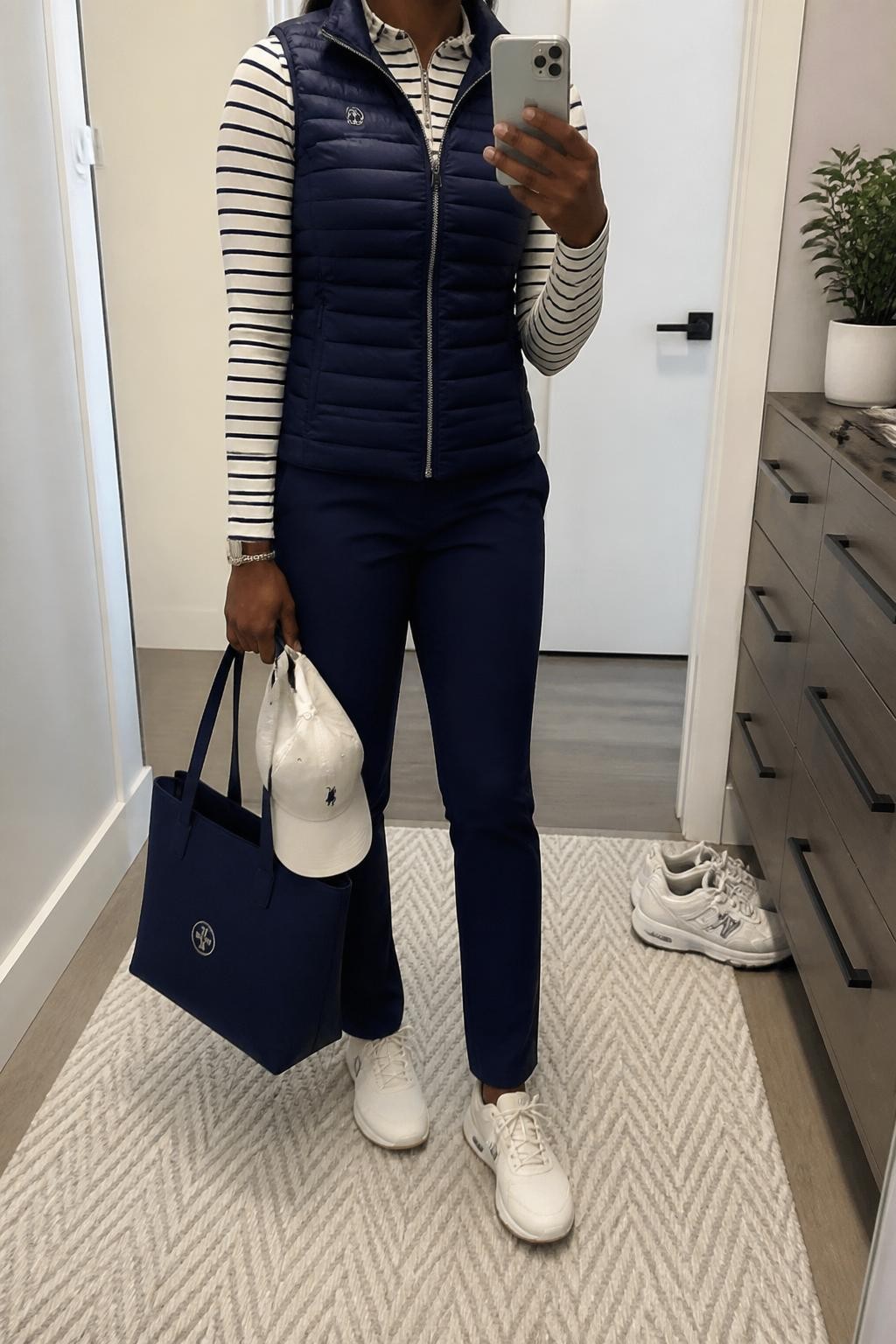 Mirror selfie of a Black woman wearing a slim navy puffer vest over a fitted cream-and-navy striped long-sleeve golf top, paired with navy straight-leg golf pants, clearly showing cream spikeless golf shoes, a cream golf cap, and a structured navy tote, holding an iPhone in front of a minimal dressing room mirror, cropped at the neck so the face does not show, relaxed casual selfie pose, soft indoor lighting, a pair of shoes on the floor nearby and a small plant on a dresser, natural unstaged setting, latest iPhone photo quality.