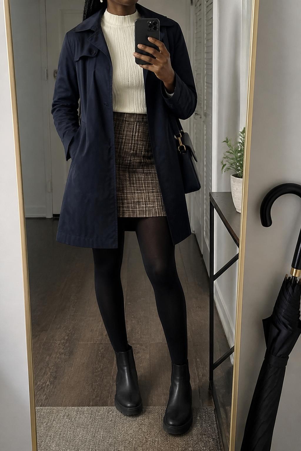mirror selfie of a Black woman wearing the exact outfit described above, clearly showing a navy water-resistant trench coat layered over a fitted ivory mock-neck sweater, brown plaid mini skirt, opaque black tights, black rubber Chelsea boots, a navy shoulder bag, and pearl stud earrings, holding a phone in front of a mirror, cropped at NECK so the face does not show, with a relaxed casual selfie pose, in a feminine apartment hallway mirror, soft gray rainy light, with an umbrella leaning beside the mirror and a small plant on a console table, latest iPhone photo quality.