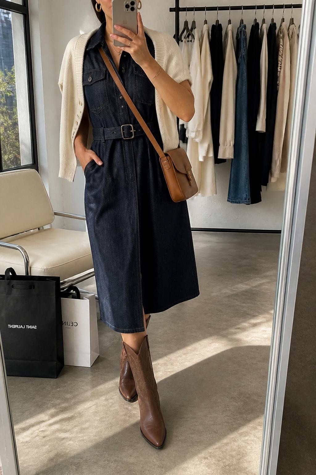 mirror selfie of a woman wearing the exact outfit described above, clearly showing brown refined western boots, a dark denim belted midi dress, a cream cropped cardigan draped over the shoulders, a tan crossbody bag, and gold hoop earrings, holding a phone in front of a mirror, cropped at NECK so the face does not show, with a relaxed casual selfie pose, in a fashion studio mirror corner, natural window light, with a clothing rack with garments behind and shopping bags beside a chair, latest iPhone photo quality.