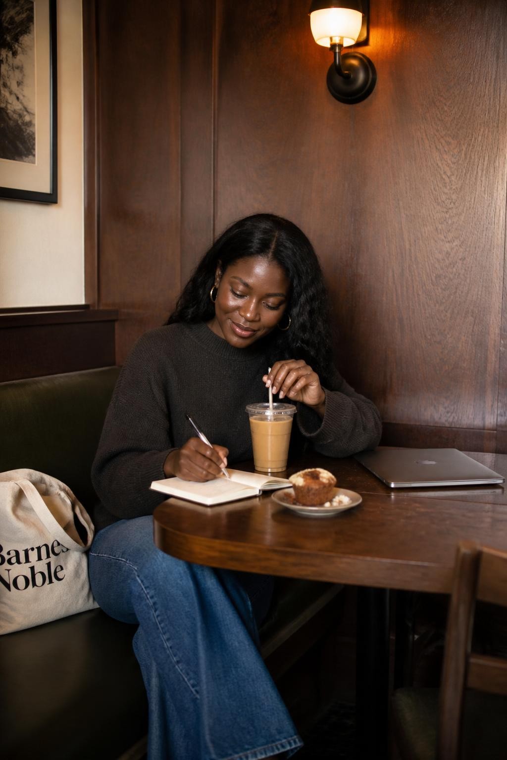 A cozy DSLR cafe photo of an adult Black woman with deep brown skin sitting alone in a corner booth, wearing an oversized charcoal sweater, wide-leg jeans, and simple hoop earrings, writing in a small notebook with one hand while holding an iced coffee straw with the other, head slightly tilted down, soft smile, laptop closed beside her, tote bag on the seat, half-eaten muffin on a plate, warm wall lamp above the booth, wood panel wall, soft shadows, realistic crumbs on the table, thoughtful cafe workday mood, natural human lifestyle photo.