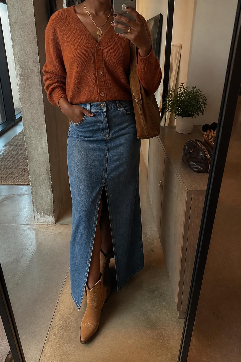 mirror selfie of a Black woman wearing the exact outfit described above, clearly showing a rust button-front cardigan tucked into a medium-wash denim maxi skirt with a front slit, tan western ankle boots, a brown suede shoulder bag, and gold layered necklaces, holding a phone in front of a mirror, cropped at NECK so the face does not show, with a relaxed casual selfie pose, in a stylish city apartment, warm moody lighting, with a small plant on a dresser and a makeup brush or cosmetic bag on the counter, latest iPhone photo quality.