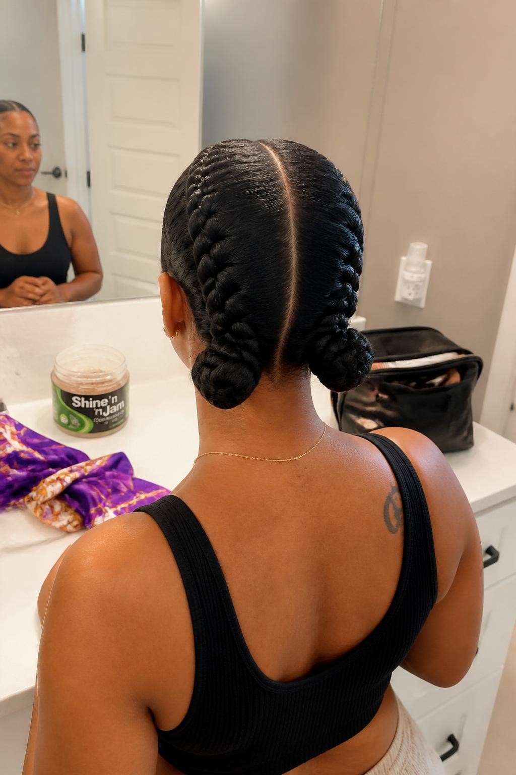 Image prompt: Photo of a Black woman wearing two smooth feed-in cornrows that finish in matching braided low buns, back-of-head view to highlight the details, bathroom vanity mirror with a makeup bag nearby, soft indoor lighting, containing an open jar of braiding gel on the counter and a silk scarf beside the sink, realistic morning styling moment, latest iPhone photo quality.