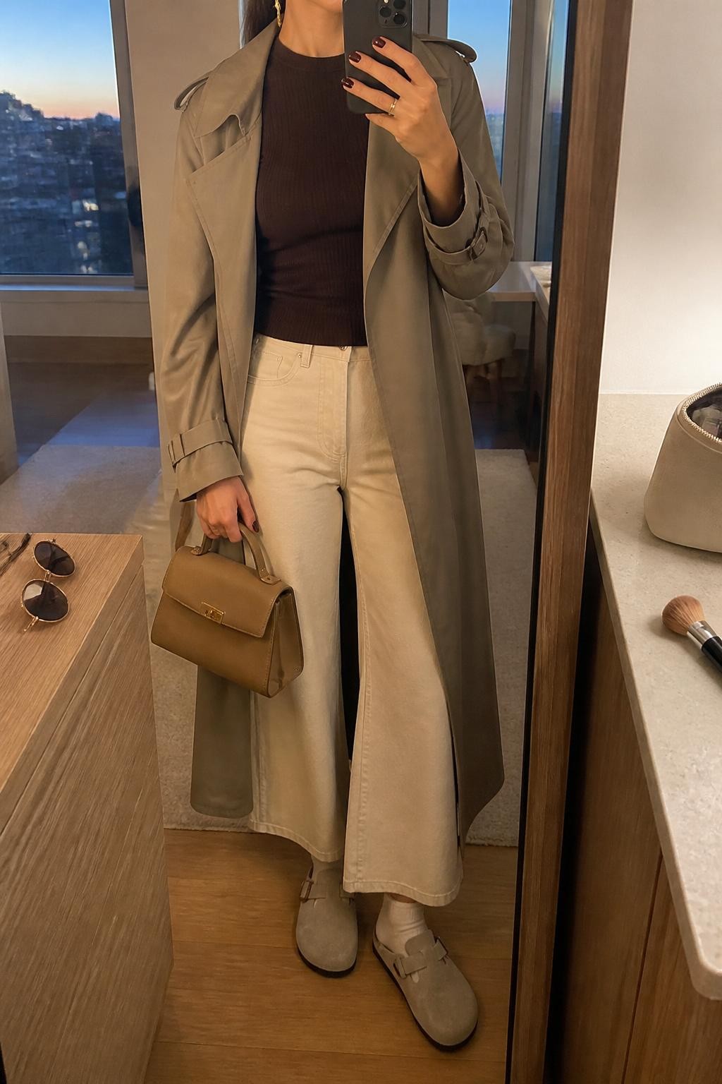 mirror selfie of a woman wearing the exact outfit described above, clearly showing stone beige suede Birkenstock-style clogs with white socks, cream wide-leg pants, a chocolate brown fitted ribbed top, a taupe trench coat worn open, a tan top-handle bag, and gold drop earrings, holding a phone in front of a mirror, cropped at NECK so the face does not show, with a relaxed casual selfie pose, in a stylish city apartment, evening golden hour, with a makeup brush or cosmetic bag on the counter and sunglasses placed on a dresser, latest iPhone photo quality.