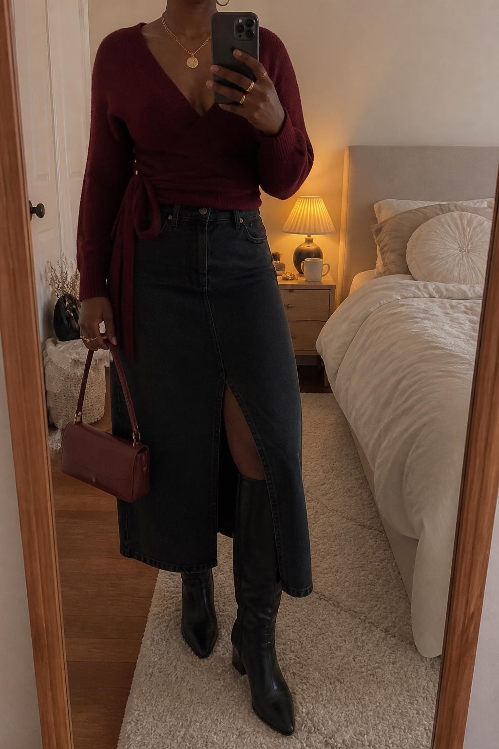 mirror selfie of a Black woman wearing the exact outfit described above, clearly showing a black denim maxi skirt with a front slit, burgundy wrap sweater, black knee-high boots, burgundy mini handbag, gold layered necklaces, and small gold hoop earrings, holding a phone in front of a mirror, cropped at NECK so the face does not show, with a relaxed casual selfie pose, in a cozy bedroom mirror setup, warm moody lighting, with a coffee mug on a bedside table and soft pillows on an unmade bed, latest iPhone photo quality.