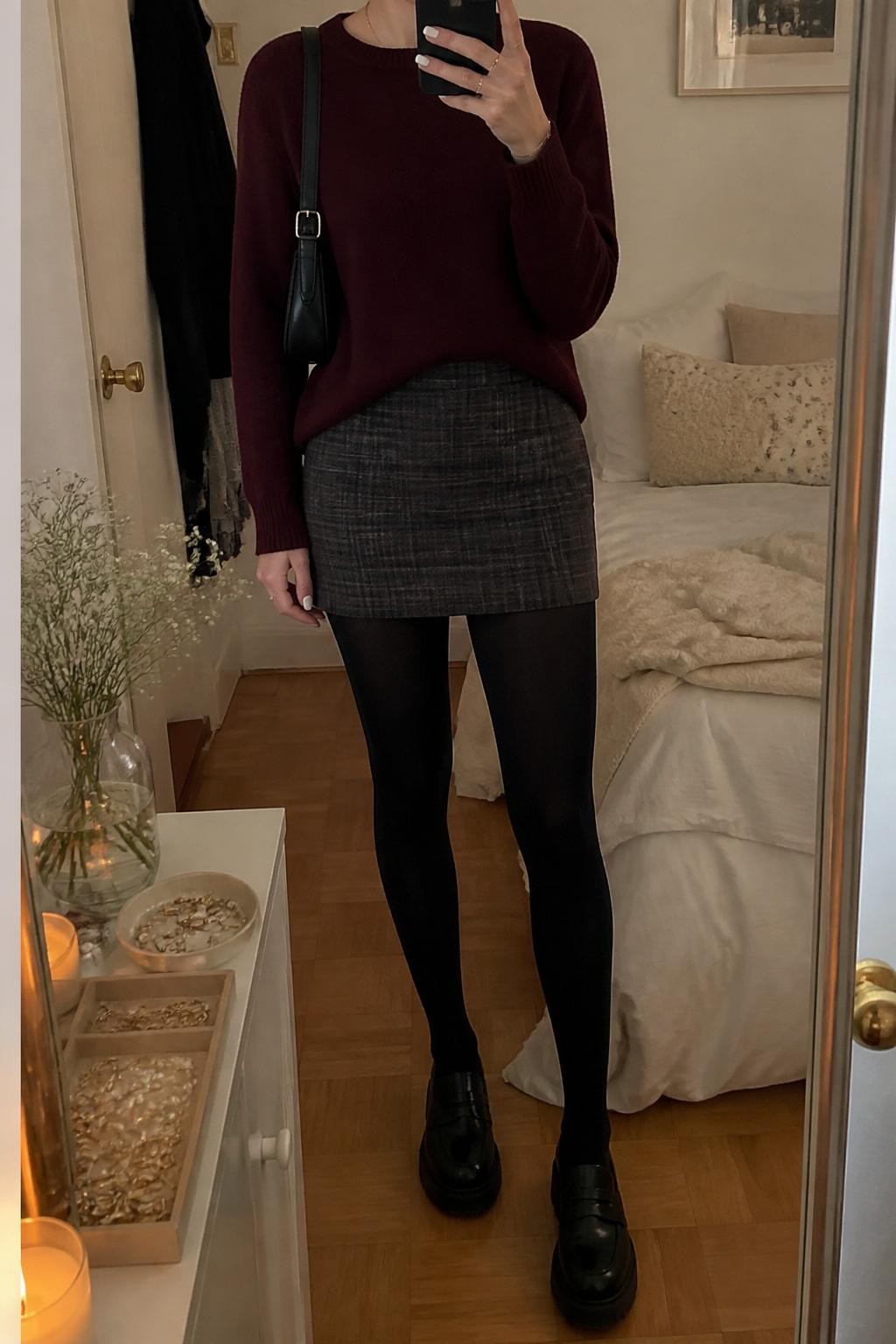 Mirror selfie of a woman wearing a burgundy crewneck sweater, gray plaid wool mini skirt, black opaque tights, black chunky loafers, pearl stud earrings, and a small black shoulder bag, holding a phone in front of a mirror, cropped at NECK so the face does not show, relaxed casual selfie pose, in a cozy bedroom mirror setup, warm indoor lighting, with soft pillows on an unmade bed and a jewelry tray on the vanity, latest iPhone photo quality.