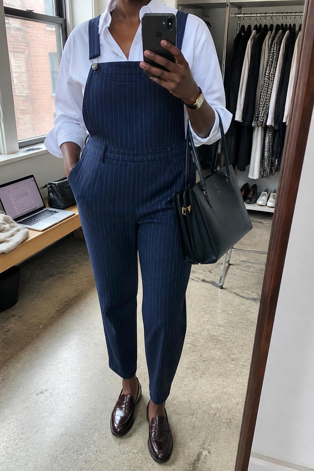 Mirror selfie of a Black woman wearing navy pinstripe tailored overalls with tapered trousers over a crisp white cotton button-down shirt, clearly showing oxblood leather loafers, a structured navy work tote, and a gold rectangular wristwatch, holding an iPhone in front of a fashion studio mirror corner, cropped at the neck so the face does not show, relaxed casual selfie pose, natural window light, an open closet with clothes visible and a laptop on a bench, natural unstaged setting, latest iPhone photo quality.