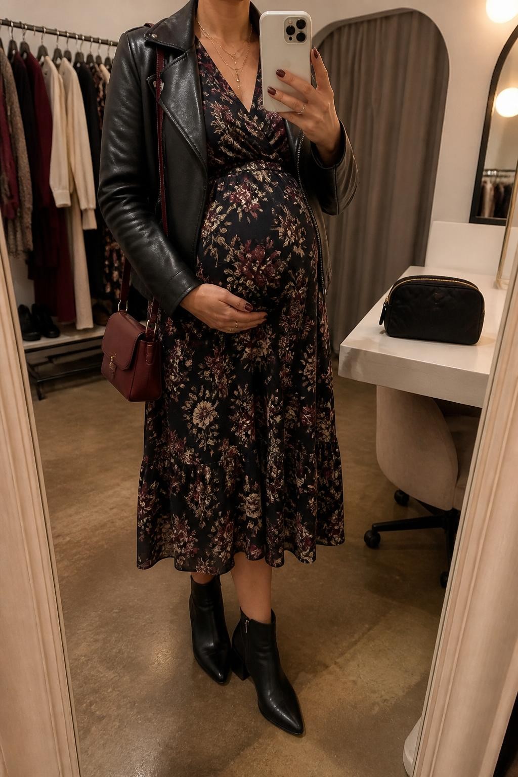 mirror selfie of a pregnant woman wearing a dark floral maternity midi dress with burgundy and cream print, cropped black leather jacket, black ankle boots, gold layered necklace, and a small burgundy handbag, clearly showing the same clothing items, colors, fabrics, and accessories listed in the Outfit Pieces section, holding a phone in front of a mirror, cropped at NECK so the face does not show, with a relaxed casual selfie pose, in a fashion studio mirror corner, warm moody lighting, including clothing rack with garments behind and a cosmetic bag on the counter, latest iPhone photo quality.