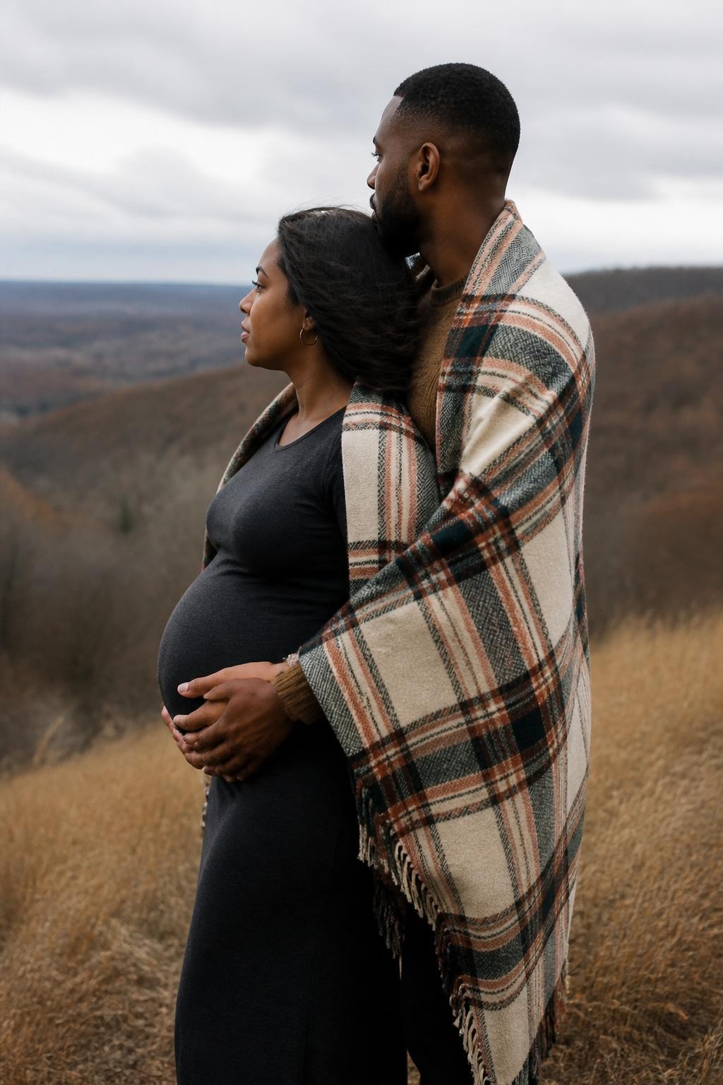 Image Prompt: A pregnant Black woman and her partner standing together on a breezy hill covered with dry fall grass, both loosely wrapped inside one oversized cream, rust, and dark-green plaid wool blanket, the woman wearing a fitted charcoal maternity dress and the partner wearing black trousers with a brown knit sweater, her back resting gently against his chest while his hands are placed naturally beneath hers around the lower curve of the bump, both looking toward the distant valley rather than the camera, cloudy sky and late-season trees behind them, photographed with a DSLR 70mm lens at chest height, realistic dark skin texture, natural blanket folds, believable wind movement, quiet expressions, no dramatic flying fabric, no mountain fantasy background.
