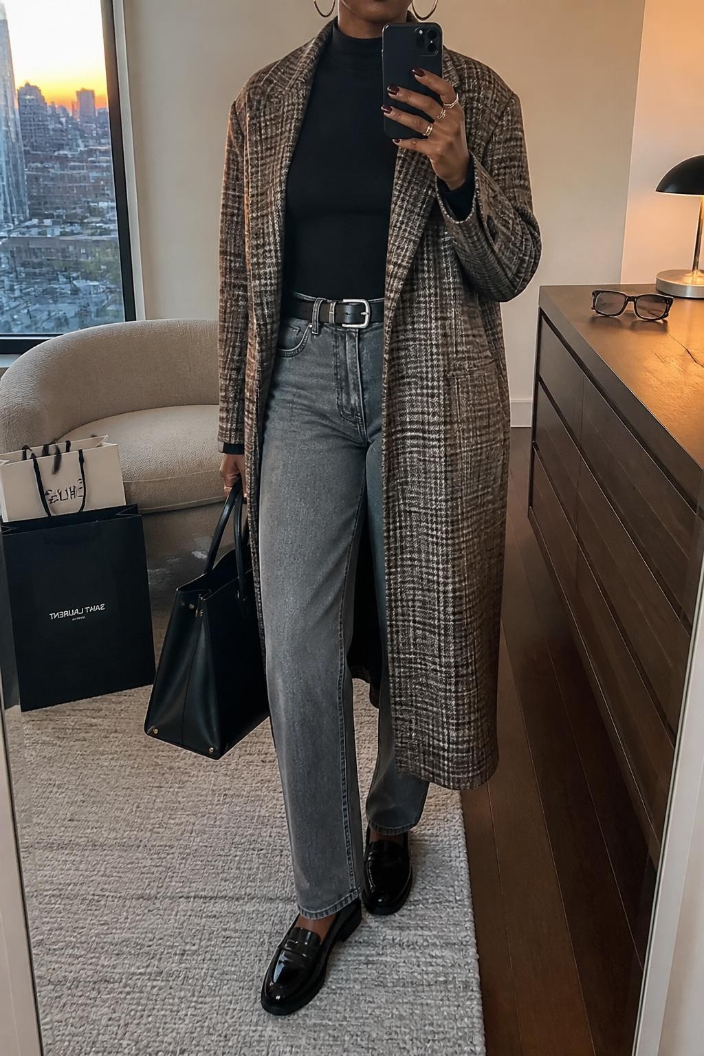 mirror selfie of a Black woman wearing the exact outfit described above, clearly showing gray straight-leg jeans, a black fitted mock-neck top, a brown and gray plaid longline coat, black loafers, a black structured tote bag, and silver hoop earrings, holding a phone in front of a mirror, cropped at NECK so the face does not show, with a relaxed casual selfie pose, in a stylish city apartment, evening golden hour, with shopping bags beside a chair and sunglasses placed on a dresser, latest iPhone photo quality.