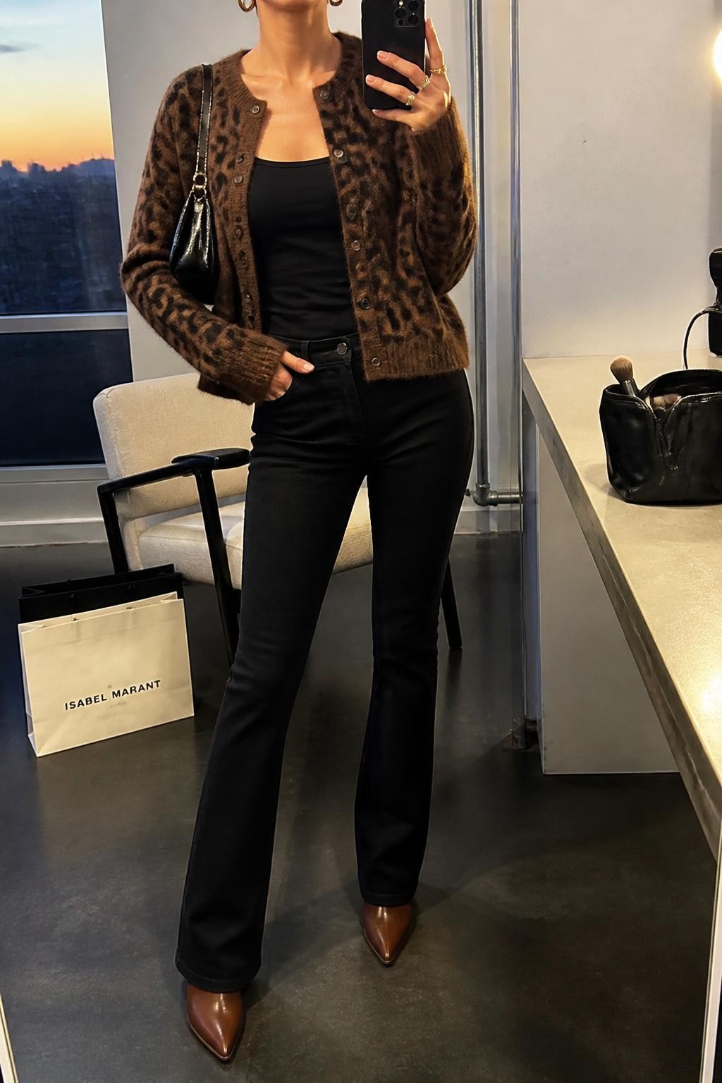 mirror selfie of a woman wearing the exact outfit described above, clearly showing black bootcut jeans, a brown leopard knit cardigan, a fitted black tank top, brown pointed ankle boots, a glossy black shoulder bag, and gold hoop earrings, holding a phone in front of a mirror, cropped at NECK so the face does not show, with a relaxed casual selfie pose, in a fashion studio mirror corner, evening golden hour, with shopping bags beside a chair and a makeup brush or cosmetic bag on the counter, latest iPhone photo quality.