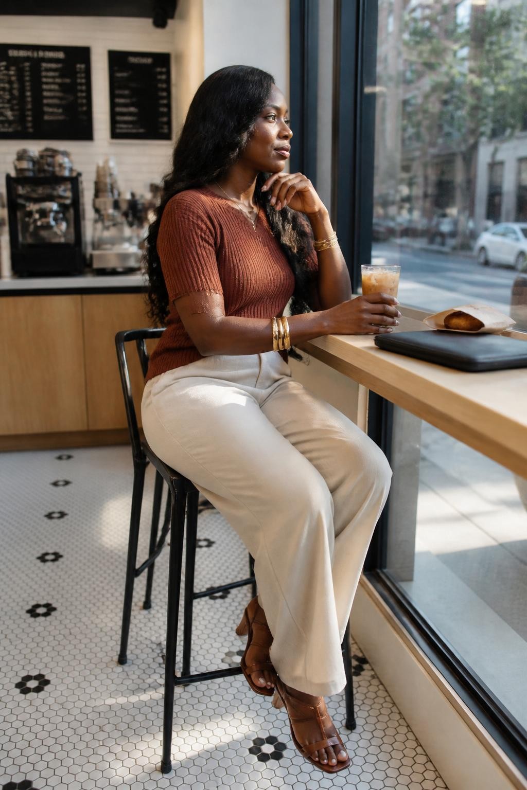 A modern cafe photo of an adult Black woman with deep brown skin sitting on a tall bar stool by a window counter, wearing a rust-colored knit top, cream wide-leg trousers, and gold bangles, body turned in side profile, one elbow resting on the counter, one hand holding a glass of iced coffee, legs crossed at the ankles, looking out through the window, city street softly blurred outside, sunlight on her cheekbone, laptop sleeve and small pastry bag on the counter, realistic reflections on the glass, clean DSLR lifestyle photo, calm city cafe mood.