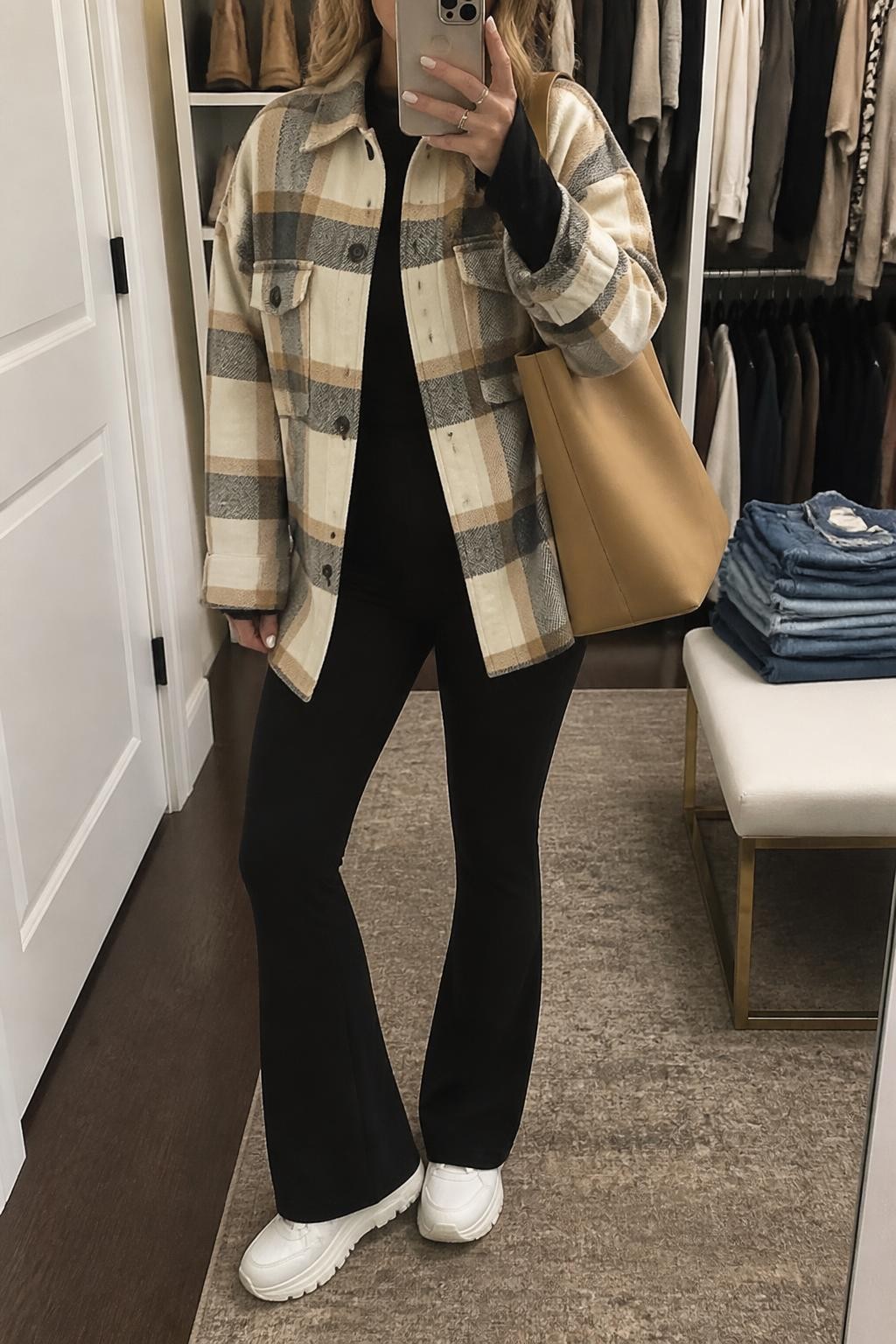 Mirror selfie of a woman wearing an oversized cream, camel, and charcoal plaid wool-blend shacket, fitted black long-sleeve top, black flared leggings, chunky white leather sneakers, and a camel canvas tote bag, clearly showing the exact clothing items, colors, fabrics, and accessories listed, holding a phone in front of a walk-in closet mirror, cropped at the neck so the face does not show, relaxed casual selfie pose, soft indoor lighting, an open closet with clothes visible and folded denim jeans on a bench, casual and uncluttered, latest iPhone photo quality.
