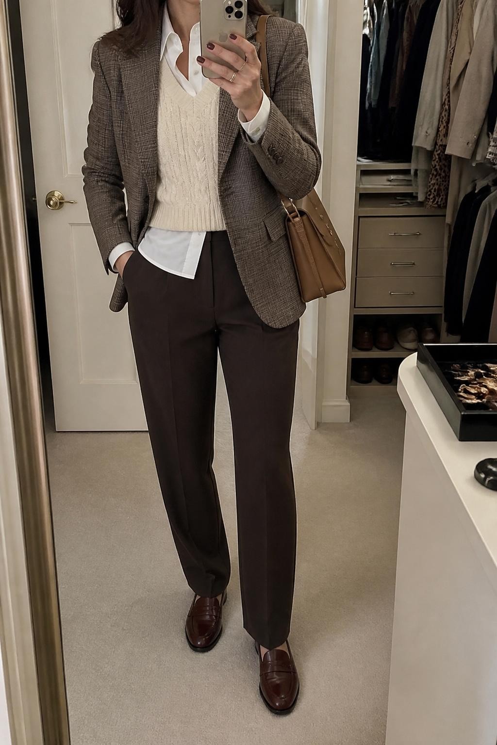 Mirror selfie of a woman wearing a tailored brown plaid blazer over a cream cable-knit sweater vest and a crisp white button-down shirt, clearly showing chocolate brown tailored trousers, burgundy leather loafers, and a structured tan shoulder bag, holding an iPhone in front of a walk-in closet mirror, cropped at the neck so the face does not show, relaxed casual selfie pose, soft indoor lighting, an open closet with clothes visible and a jewelry tray on the vanity, natural unstaged setting, latest iPhone photo quality.