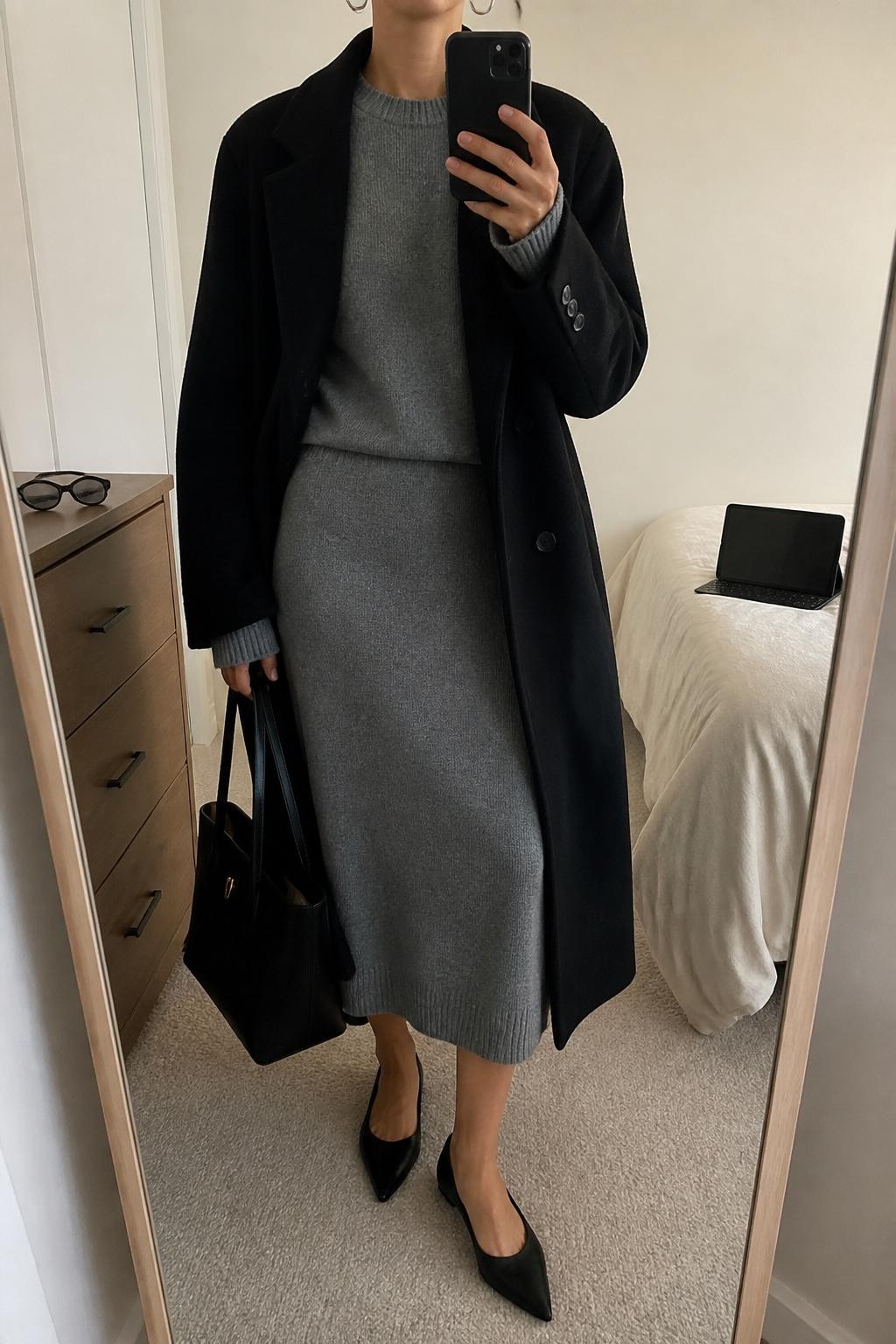 mirror selfie of a woman wearing the exact outfit described above, clearly showing a gray knit sweater, matching gray knit midi skirt, black wool coat worn open, black pointed flats, a structured black tote bag, and silver hoop earrings, holding a phone in front of a mirror, cropped at NECK so the face does not show, with a relaxed casual selfie pose, in a minimal dressing room, soft indoor lighting, with a laptop or tablet on the bed and sunglasses placed on a dresser, latest iPhone photo quality.