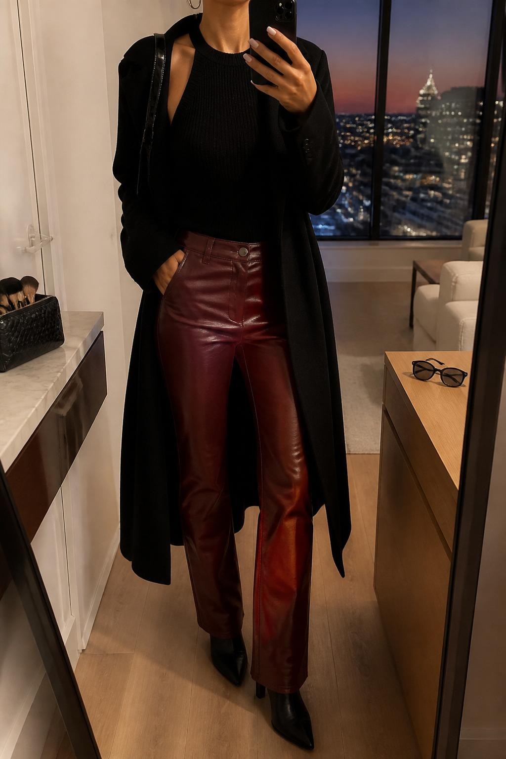 mirror selfie of a woman wearing the exact outfit described above, clearly showing red leather straight-leg pants, a black knit halter top, black pointed ankle boots, a black longline coat, a glossy black shoulder bag, and silver hoop earrings, holding a phone in front of a mirror, cropped at NECK so the face does not show, with a relaxed casual selfie pose, in a stylish city apartment, evening golden hour, with a makeup brush or cosmetic bag on the counter and sunglasses placed on a dresser, latest iPhone photo quality.