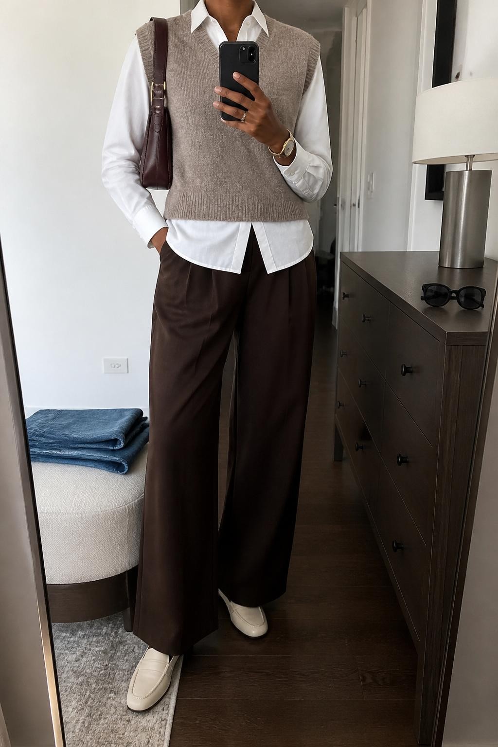 mirror selfie of a Black woman wearing the exact outfit described above, clearly showing a taupe sweater vest layered over a crisp white poplin shirt, chocolate brown wide-leg trousers, cream loafers, a burgundy shoulder bag, and a slim gold watch, holding a phone in front of a mirror, cropped at NECK so the face does not show, with a relaxed casual selfie pose, in a stylish city apartment, soft indoor lighting, with sunglasses placed on a dresser and folded denim jeans on a bench, latest iPhone photo quality.