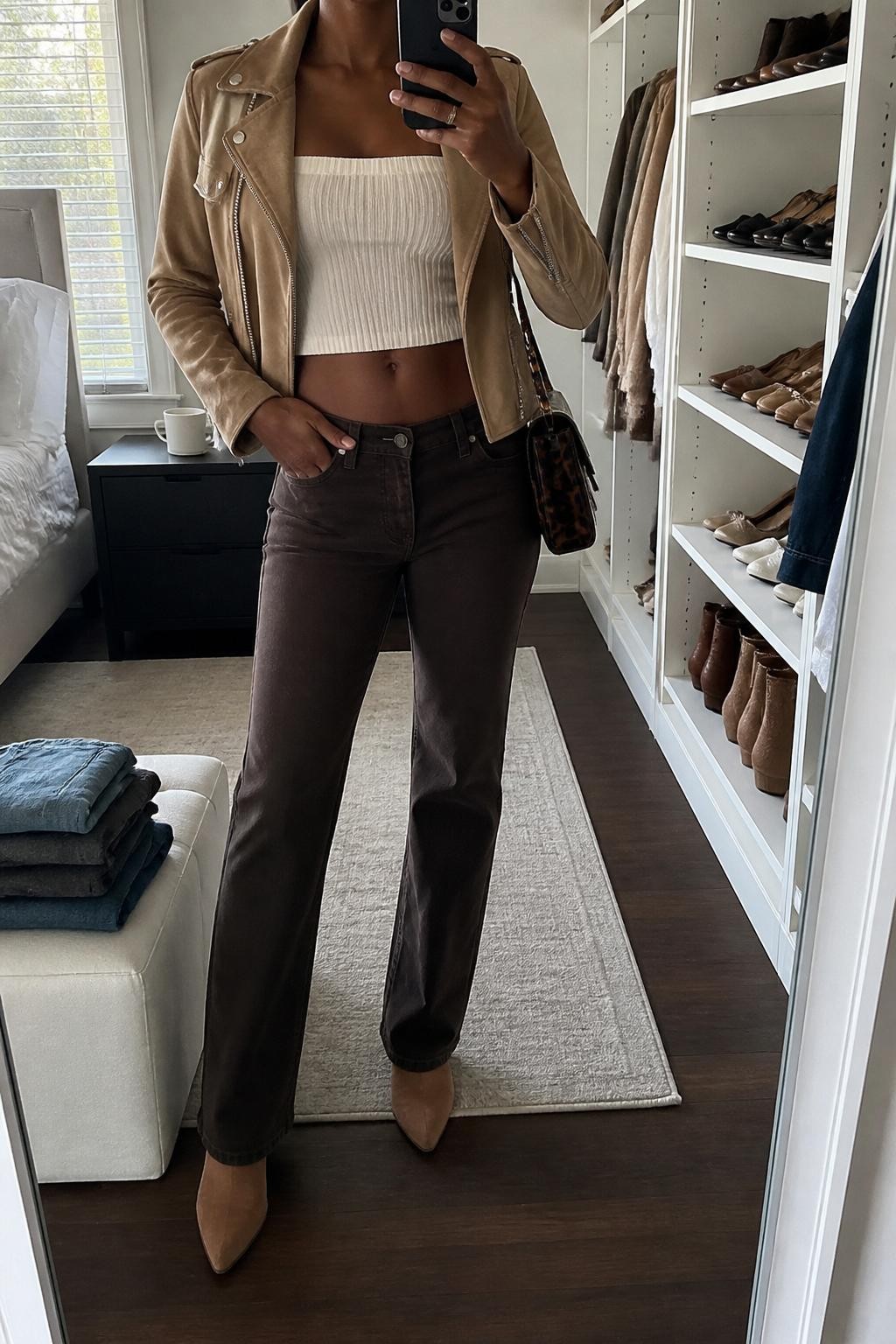 mirror selfie of a Black woman wearing a tan suede moto jacket, cream knit tube top, dark brown low-rise straight jeans, tan pointed ankle boots, and a tortoiseshell mini shoulder bag, clearly showing the same clothing items, colors, fabrics, and accessories listed in the Outfit Pieces section, holding a phone in front of a mirror, cropped at NECK so the face does not show, with a relaxed casual selfie pose, in a walk-in closet mirror, natural window light, including folded denim jeans on a bench and a coffee mug on a bedside table, latest iPhone photo quality.