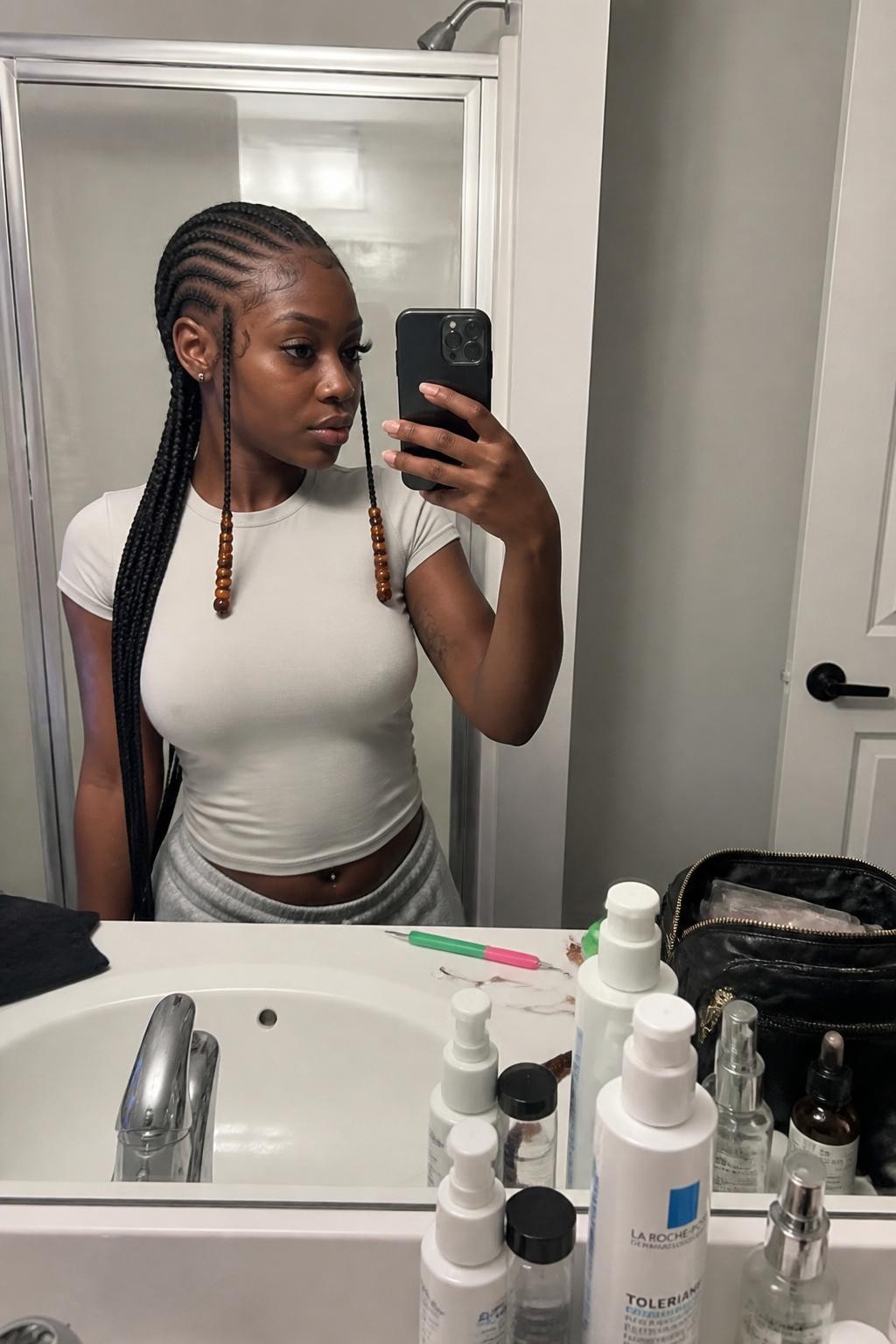 Image prompt: Photo of a Black woman wearing detailed Fulani braids with a center cornrow, curved side braids, and amber beads near the face, candid mirror selfie, bathroom vanity mirror with skincare bottles nearby, soft indoor lighting, containing a beading tool on the counter and a makeup bag beside the sink, realistic styling moment, latest iPhone photo quality.