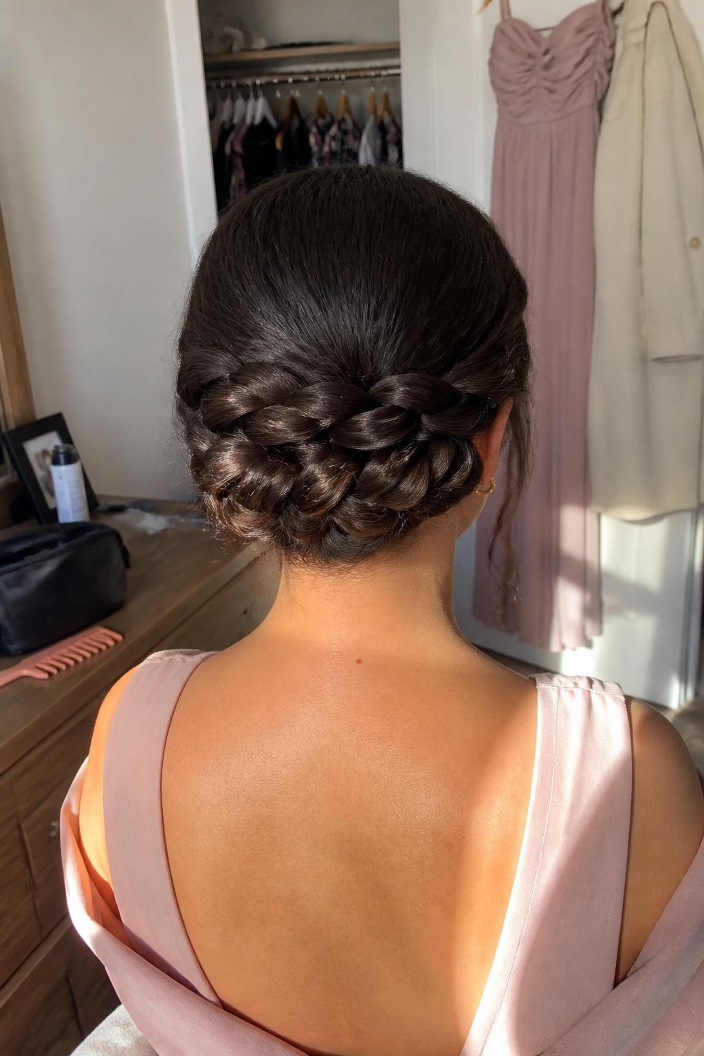 Image prompt: Photo of a woman wearing a modern rope-braid updo made from three thick twisted loops arranged across the nape, back-of-head view to highlight the detailed shape, sun-drenched bedroom with a formal dress and coat visible near an open closet, natural window light, containing a tail comb on the dresser and a makeup bag nearby, realistic fall wedding guest preparation, latest iPhone photo quality.