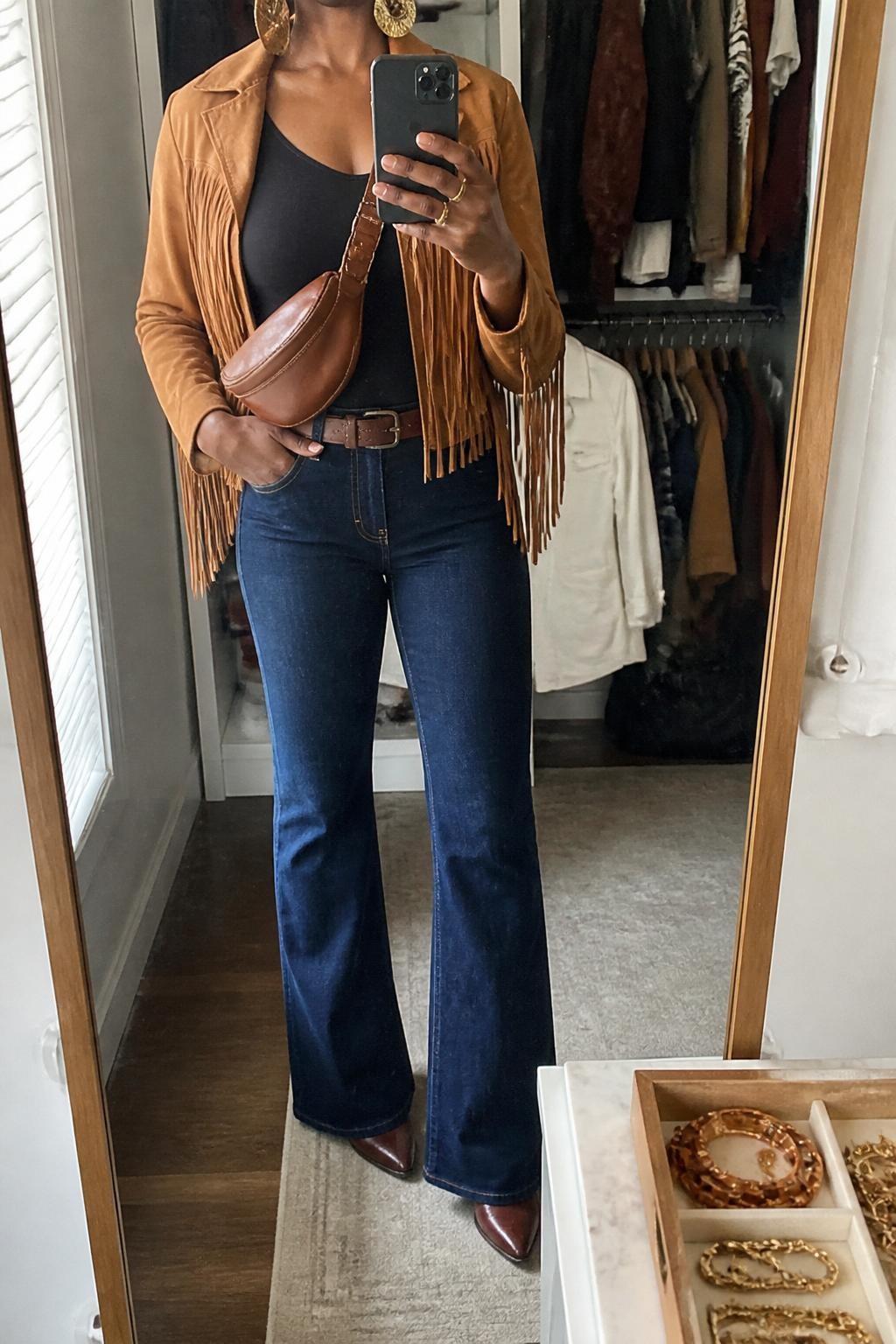 mirror selfie of a Black woman wearing the exact outfit described above, clearly showing a tan suede fringe jacket layered over a fitted black bodysuit, dark blue flare jeans, brown western boots, a cognac belt bag worn crossbody, and gold statement earrings, holding a phone in front of a mirror, cropped at NECK so the face does not show, with a relaxed casual selfie pose, in a walk-in closet mirror, natural window light, with an open closet with clothes visible and a jewelry tray on the vanity, latest iPhone photo quality.