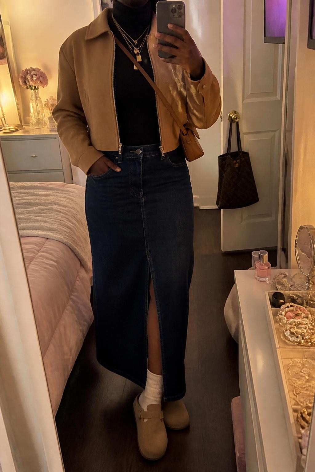mirror selfie of a Black woman wearing the exact outfit described above, clearly showing camel suede Birkenstock-style clogs with cream socks, a dark denim maxi skirt with a front slit, a fitted black turtleneck, a camel cropped jacket, a tan crossbody bag, and gold layered necklaces, holding a phone in front of a mirror, cropped at NECK so the face does not show, with a relaxed casual selfie pose, in a feminine-themed bedroom, evening golden hour, with a jewelry tray on the vanity and a handbag hanging on a door handle, latest iPhone photo quality.
