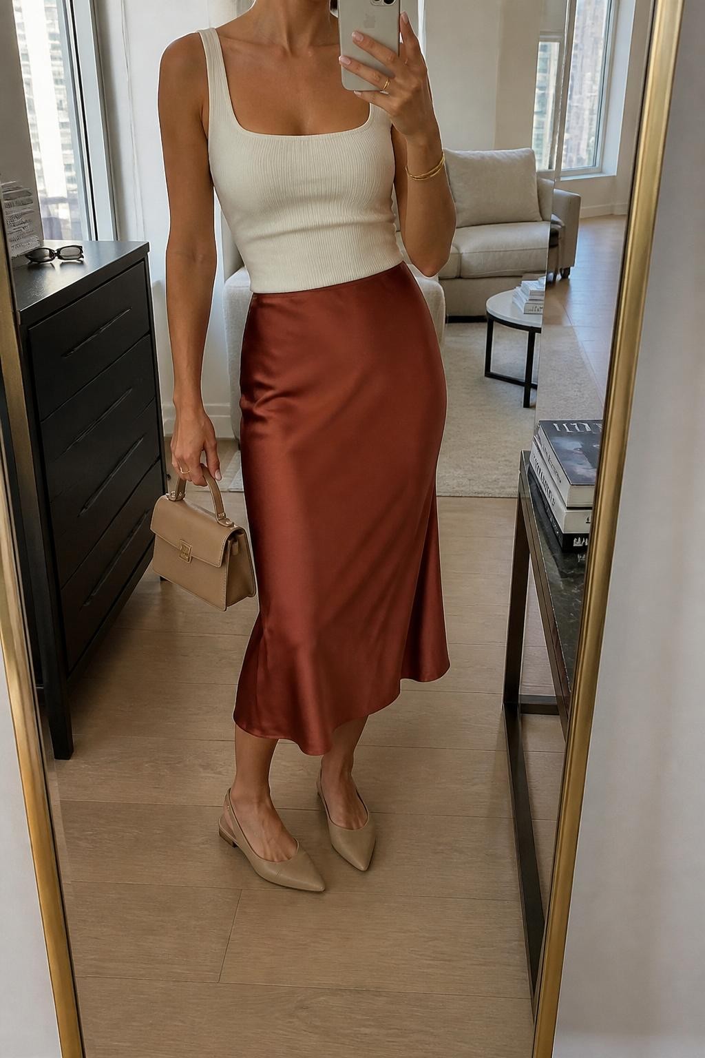 mirror selfie of a woman wearing the exact outfit described above, clearly showing a rust satin midi skirt, fitted ivory square-neck tank top, nude slingback flats, a small tan top-handle bag, and gold bangle bracelet, holding a phone in front of a mirror, cropped at NECK so the face does not show, with a relaxed casual selfie pose, in a stylish city apartment, soft indoor lighting, with sunglasses placed on a dresser and fashion magazines stacked on a side table, latest iPhone photo quality.
