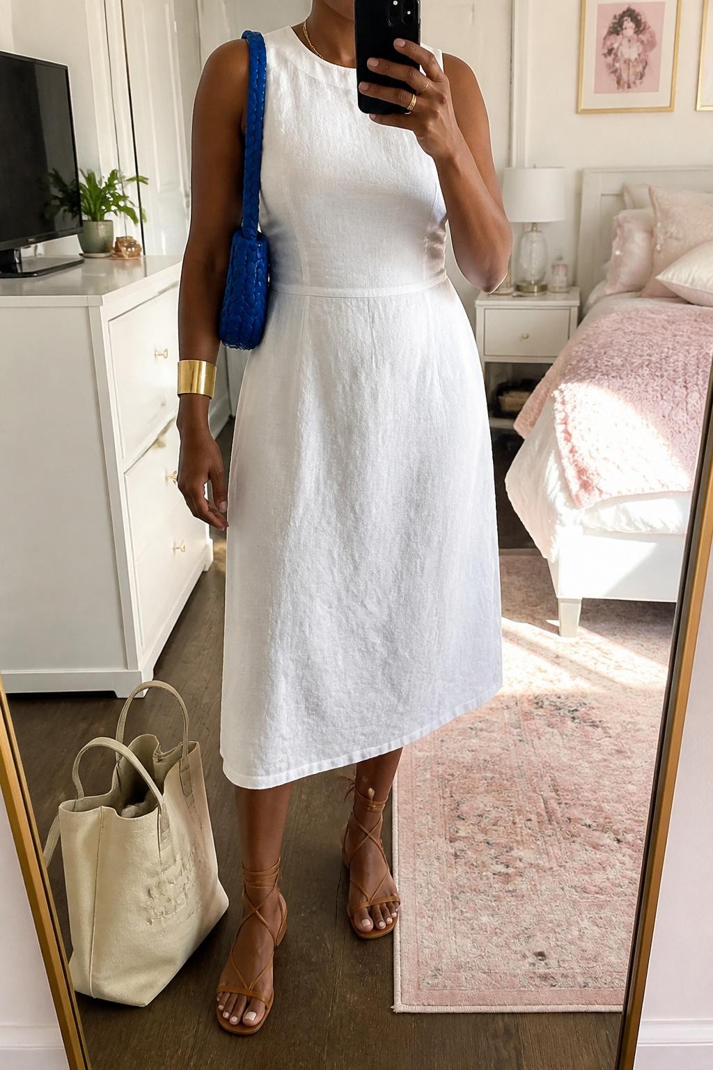 mirror selfie of a Black woman wearing a white sleeveless linen midi dress with a gently fitted waist, tan lace-up flat sandals, cobalt blue woven shoulder bag, gold cuff bracelet, and small gold hoop earrings, clearly showing the same clothing items, colors, fabrics, and accessories listed in the Outfit Pieces section, holding a phone in front of a mirror, cropped at NECK so the face does not show, with a relaxed casual selfie pose, in a feminine-themed bedroom, natural window light, including a cream tote bag resting on the floor and a small plant on a dresser, latest iPhone photo quality.