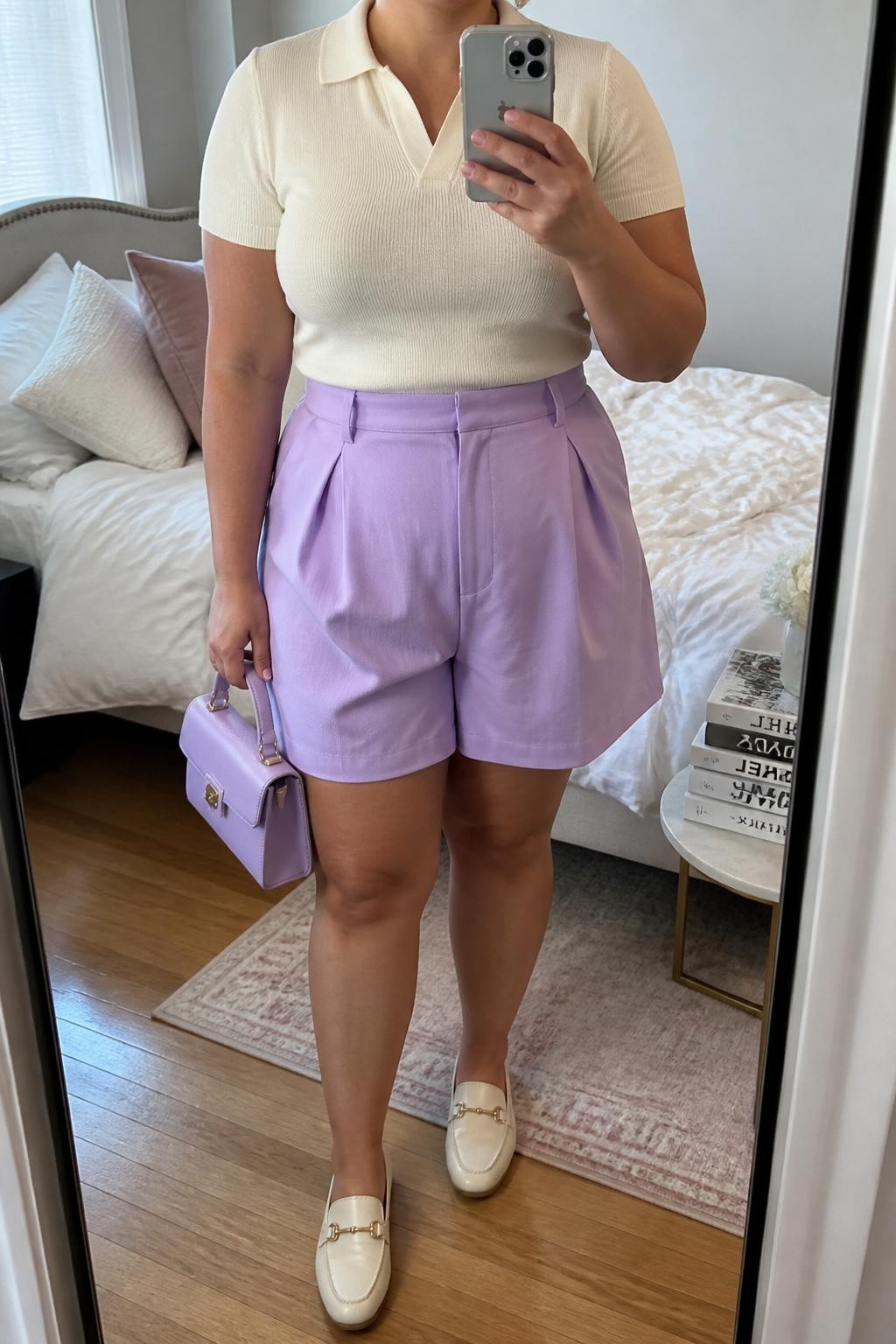 Mirror selfie of a plus size woman wearing lavender high-waisted pleated shorts with a relaxed tailored fit, a fitted cream short-sleeve knit polo, cream leather loafers, a small lavender top-handle bag, and pearl stud earrings, clearly showing the exact clothing items, lavender fabric, cream knit, pleats, footwear, and accessories listed, holding a phone in front of a mirror, cropped at the neck so the face does not appear, relaxed soft selfie pose, in a cozy bedroom mirror setup with soft indoor lighting, soft pillows on an unmade bed and a stack of fashion magazines on a side table, realistic feminine summer styling, latest iPhone photo quality.
