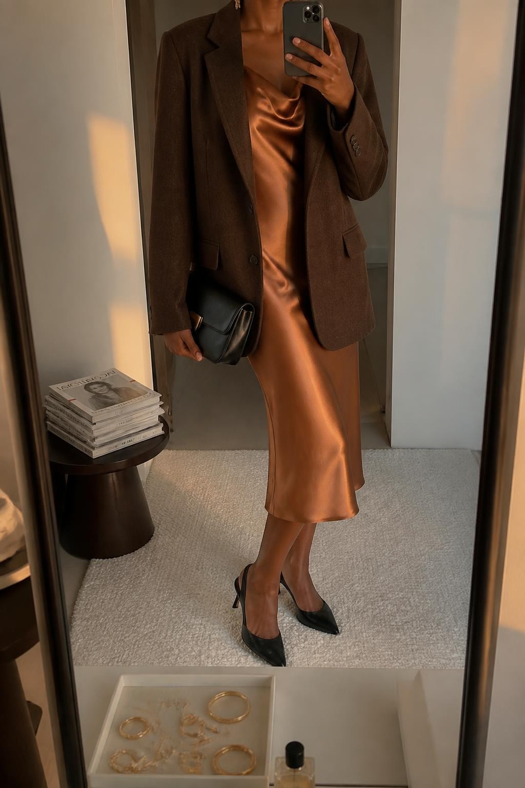 Mirror selfie of a Black woman wearing a copper satin midi slip dress, oversized cocoa-brown wool blazer, black leather slingback heels, a structured black clutch, and delicate gold drop earrings, clearly showing the exact clothing items, colors, fabrics, and accessories listed, holding a phone in front of a minimal dressing room mirror, cropped at the neck so the face does not show, relaxed casual selfie pose, evening golden-hour lighting, a jewelry tray on the vanity and a stack of fashion magazines on a side table, elegant natural setting, latest iPhone photo quality.