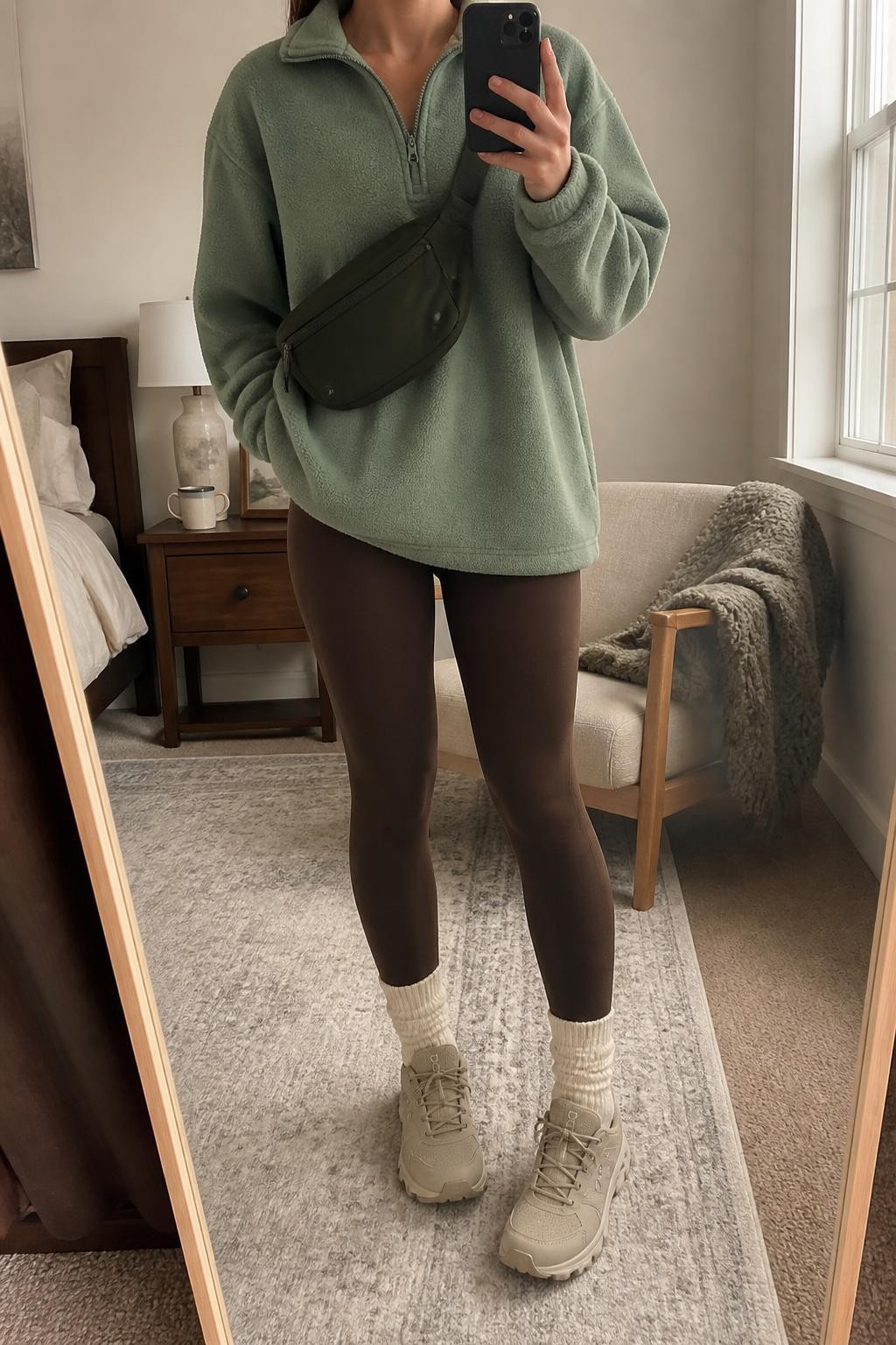 Mirror selfie of a woman wearing an oversized sage-green half-zip fleece pullover, cocoa-brown fleece-lined leggings, cream ribbed crew socks, beige waterproof hiking sneakers, and a dark green nylon belt bag, clearly showing the exact clothing items, colors, fabrics, and accessories listed, holding a phone in front of a cozy bedroom mirror setup, cropped at the neck so the face does not show, relaxed casual selfie pose, natural window light, a coffee mug on a bedside table and a folded blanket on a chair, cozy realistic setting, latest iPhone photo quality.