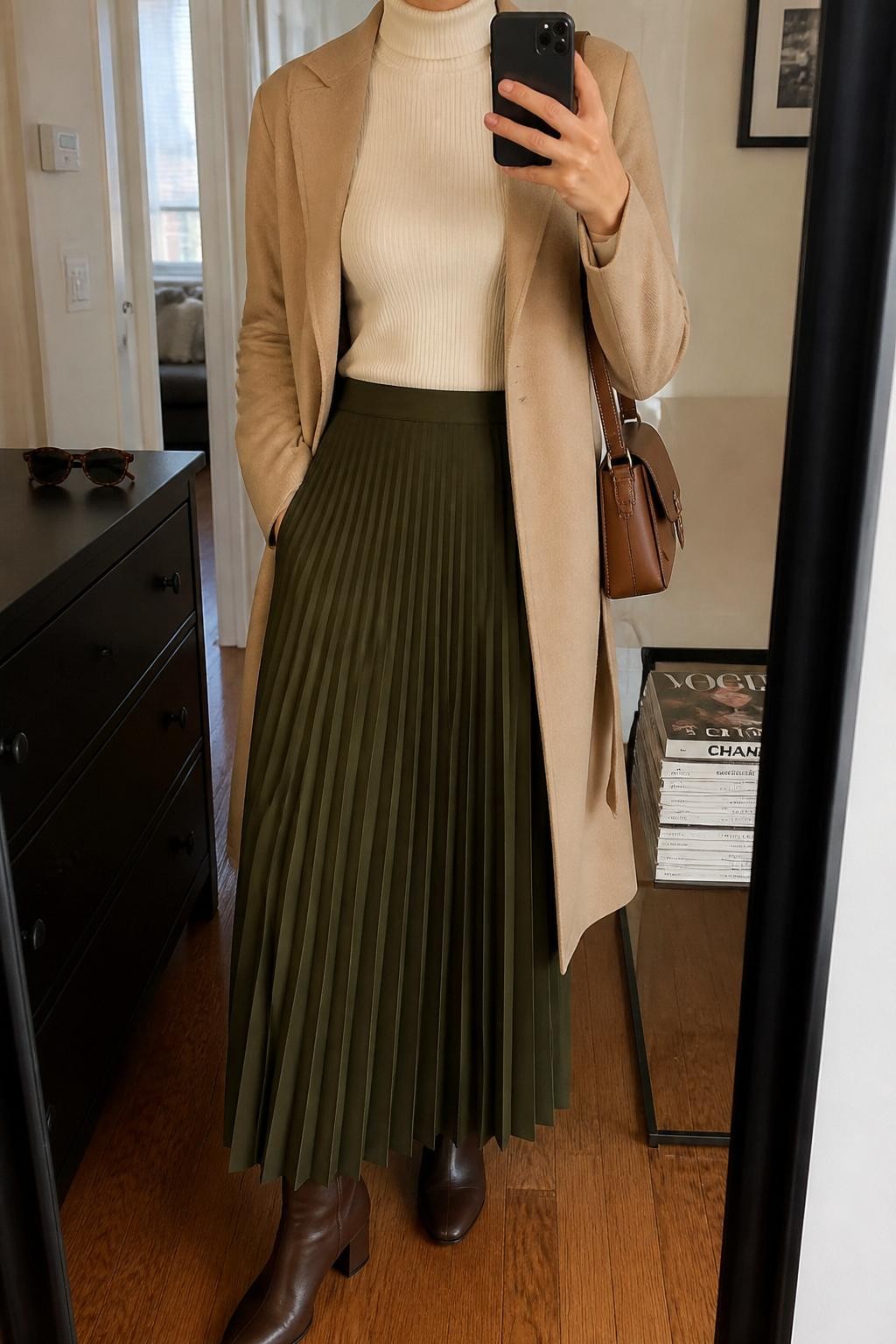 mirror selfie of a woman wearing the exact outfit described above, clearly showing an olive green pleated maxi skirt, fitted ivory turtleneck sweater, dark brown ankle boots, beige belted coat worn open, brown leather shoulder bag, and pearl stud earrings, holding a phone in front of a mirror, cropped at NECK so the face does not show, with a relaxed casual selfie pose, in a stylish city apartment, natural window light, with a stack of fashion magazines on a side table and sunglasses placed on a dresser, latest iPhone photo quality.