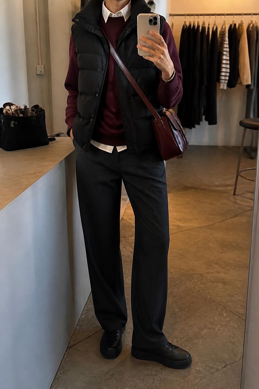 Mirror selfie of a woman wearing a black tailored puffer vest over a burgundy crew-neck sweatshirt and a white collared shirt with the collar visible, clearly showing charcoal straight-leg trousers, black leather sneakers, and a burgundy crossbody bag, holding an iPhone in front of a fashion studio mirror corner, cropped at the neck so the face does not show, relaxed casual selfie pose, warm moody lighting, a clothing rack with garments behind and a makeup bag on the counter, natural unstaged setting, latest iPhone photo quality.