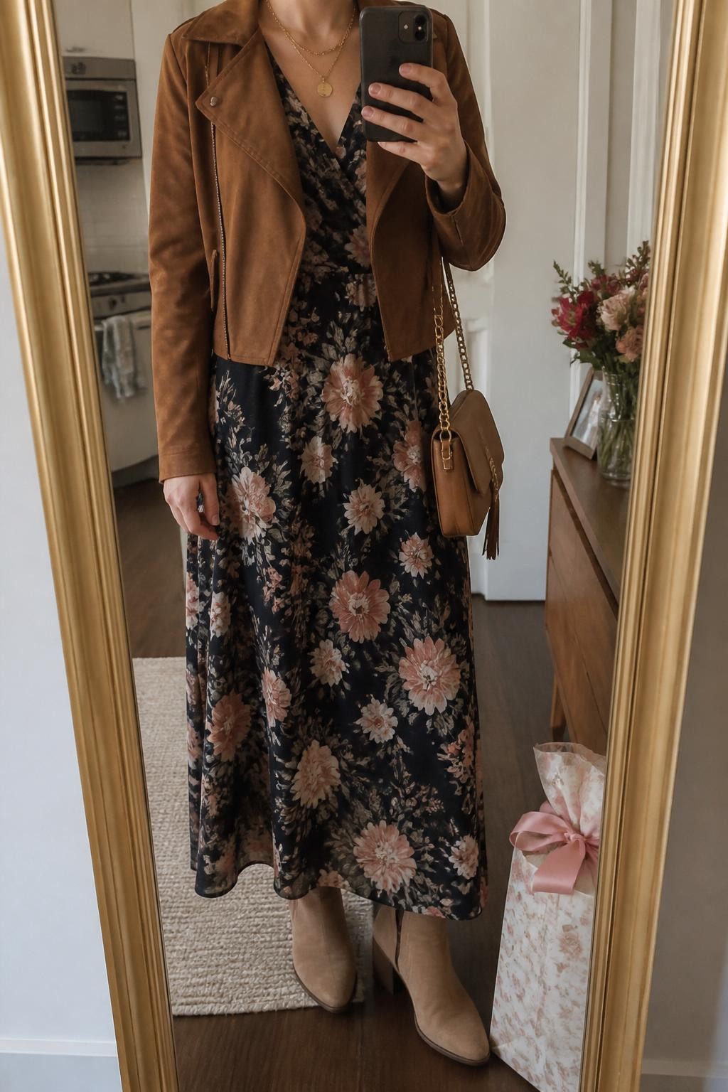 mirror selfie of a woman wearing the exact outfit described above, clearly showing a dark floral maxi dress with blush and cream flowers, brown suede cropped jacket, tan heeled ankle boots, a caramel crossbody bag, and gold layered necklaces, holding a phone in front of a mirror, cropped at NECK so the face does not show, with a relaxed casual selfie pose, in a warm apartment mirror setup, soft cloudy daylight, with a wrapped gift bag on the floor and a small bouquet on a dresser, latest iPhone photo quality.
