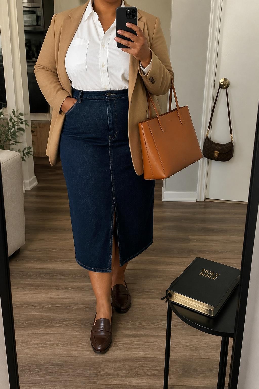 Mirror selfie of a plus size woman wearing a dark indigo high-waisted denim midi skirt with a front walking slit, a fitted white cotton button-down shirt tucked into the skirt, a camel tailored blazer worn open, dark brown leather loafers, and a structured tan tote bag, clearly showing the exact clothing items, dark denim, white cotton, camel blazer, footwear, and accessories listed, holding a phone in front of a mirror, cropped at the neck so her face does not show, relaxed polished selfie stance, in a stylish city apartment with soft indoor lighting, a Bible resting on a side table and a handbag hanging on a door handle, realistic casual church outfit, latest iPhone photo quality.