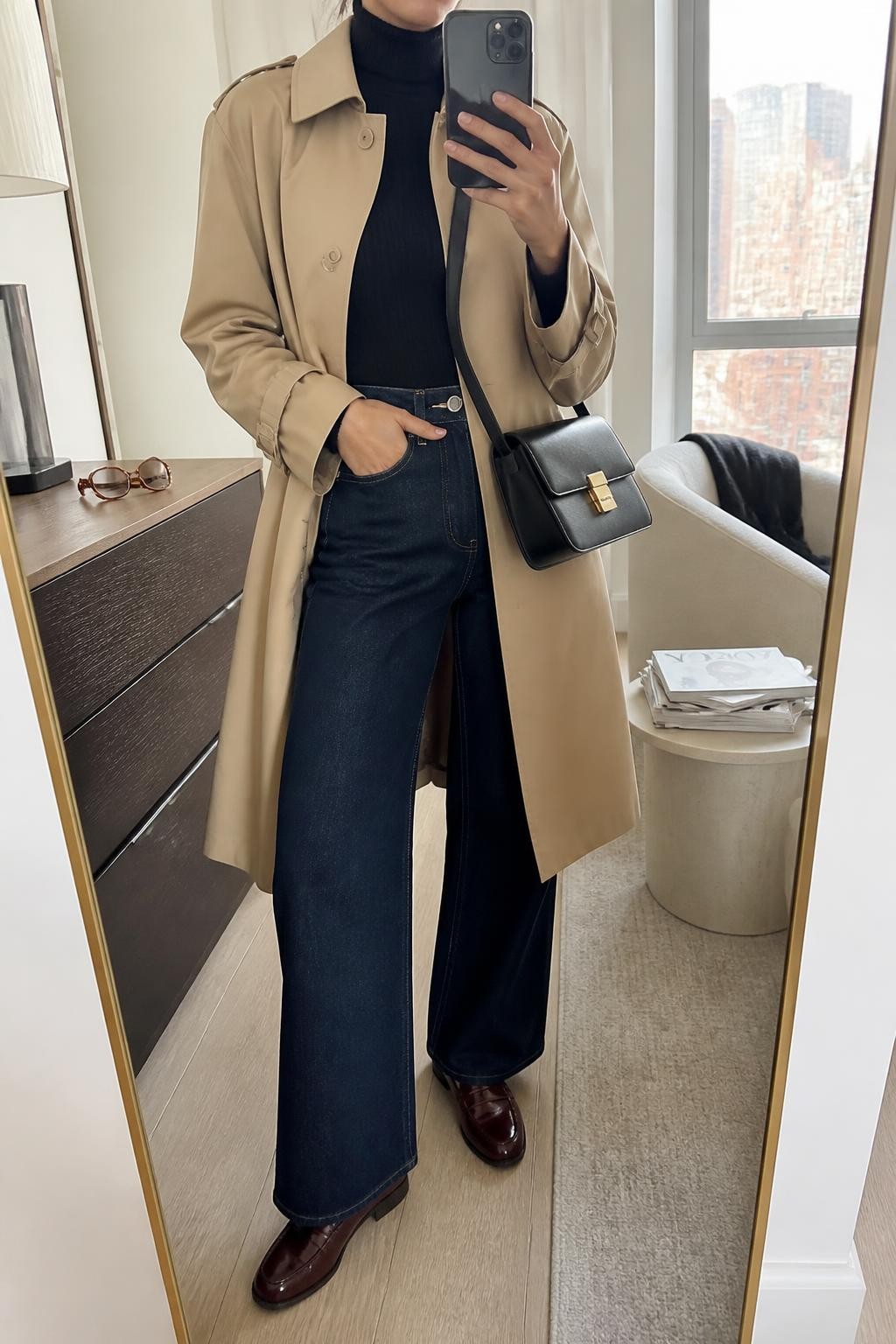 Mirror selfie of a woman wearing a knee-length camel water-resistant trench coat, fitted black ribbed turtleneck, dark indigo high-waisted wide-leg jeans, burgundy leather loafers, and a structured black crossbody bag, clearly showing the exact clothing items, colors, fabrics, and accessories listed, holding a phone in front of a stylish city apartment mirror, cropped at the neck so the face does not show, relaxed confident selfie pose, natural window light, sunglasses placed on a dresser and a stack of fashion magazines on a side table, natural and uncluttered, latest iPhone photo quality.