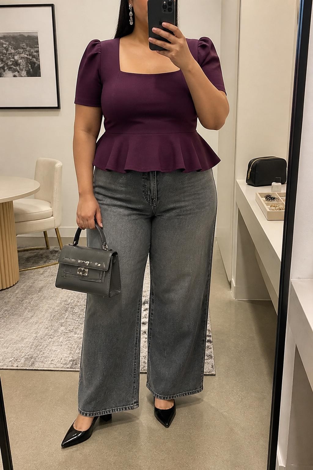 Mirror selfie of a plus size woman wearing washed grey high-waisted wide-leg jeans, a plum purple peplum top with a square neckline and short structured sleeves, black pointed block heels, a charcoal top-handle bag, and silver drop earrings, clearly showing the exact clothing items, grey denim, plum fabric, peplum shape, footwear, and accessories listed, holding a phone in front of a mirror, cropped at the neck so her face is hidden, relaxed polished selfie pose, in a fashion studio mirror corner with soft indoor lighting, a cosmetic bag on the counter and a jewelry tray on the vanity, realistic office-to-dinner styling, latest iPhone photo quality.