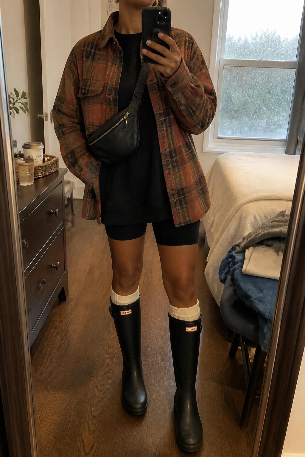 mirror selfie of a Black woman wearing the exact outfit described above, clearly showing a rust and brown plaid shacket layered over an oversized black sweatshirt, black bike shorts, black tall rain boots, cream crew socks peeking over the boots, a black belt bag worn crossbody, and small gold stud earrings, holding a phone in front of a mirror, cropped at NECK so the face does not show, with a relaxed casual selfie pose, in a cozy bedroom mirror setup, rainy morning light, with a coffee cup on a dresser and folded clothes on a chair, latest iPhone photo quality.
