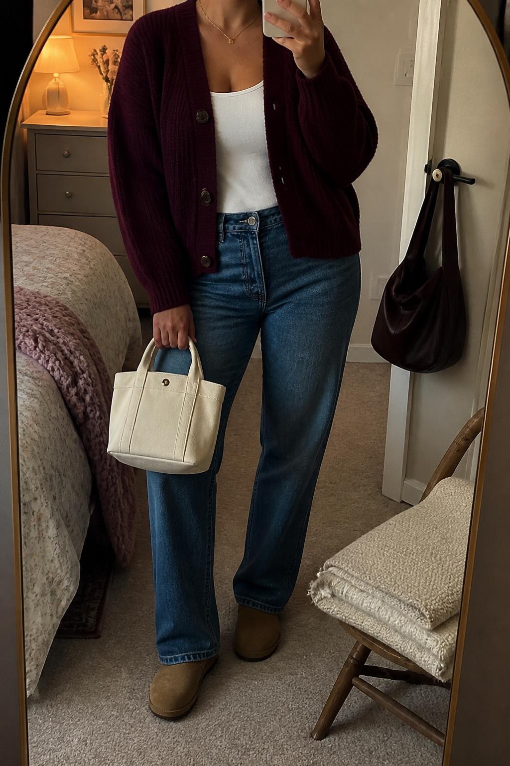 Mirror selfie of a curvy woman wearing a burgundy chunky button-front cardigan, white fitted tank top, medium-wash straight-leg jeans, tan suede clogs, and a cream mini tote bag, holding a phone in front of a mirror, cropped at NECK so the face does not show, relaxed casual selfie pose, in a feminine-themed bedroom, warm moody lighting, with a handbag hanging on a door handle and a folded blanket on a chair, latest iPhone photo quality.