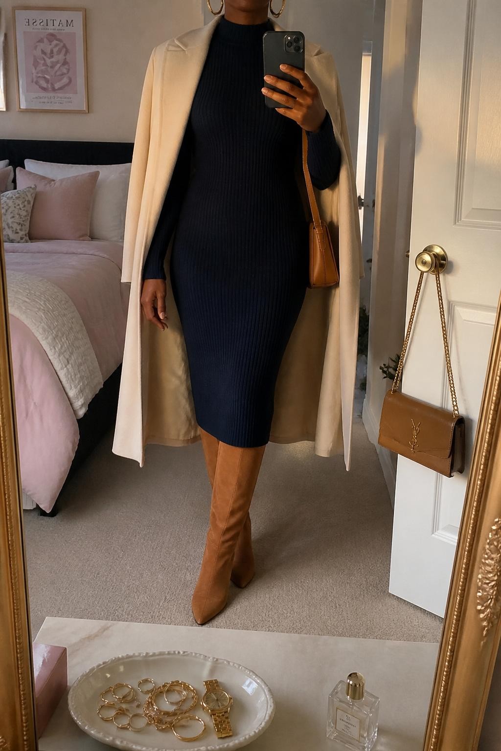 mirror selfie of a Black woman wearing the exact outfit described above, clearly showing caramel suede knee-high boots, a navy long-sleeve sweater midi dress, a cream wool coat draped open, a caramel shoulder bag, and gold hoop earrings, holding a phone in front of a mirror, cropped at NECK so the face does not show, with a relaxed casual selfie pose, in a feminine-themed bedroom, evening golden hour, with a jewelry tray on the vanity and a handbag hanging on a door handle, latest iPhone photo quality.