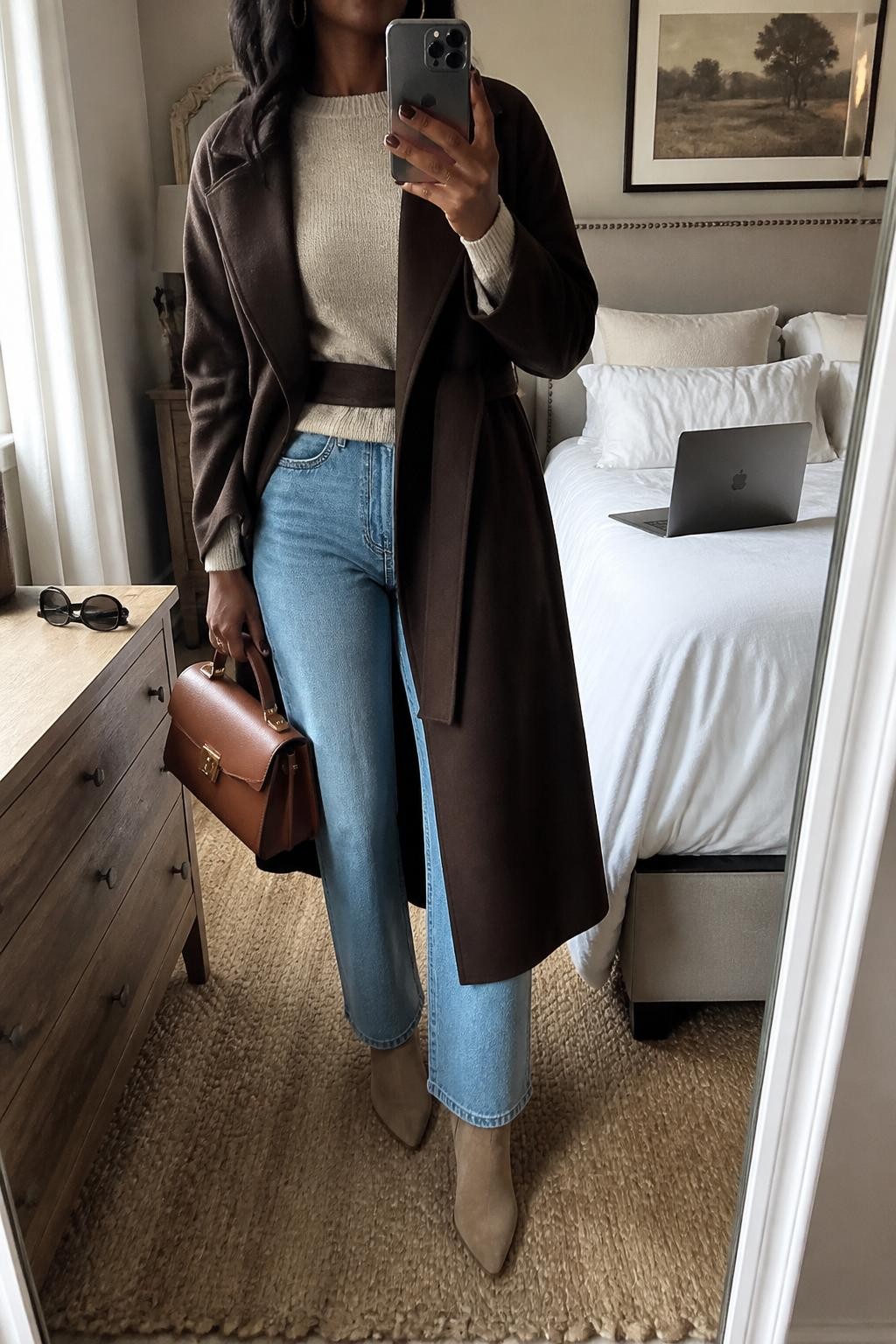 Mirror selfie of a Black woman wearing a long chocolate brown wrap coat over a fitted beige cashmere sweater and light blue straight-leg jeans, clearly showing tan suede ankle boots, a structured cognac handbag, and delicate gold hoop earrings, holding an iPhone in front of a cozy bedroom mirror setup, cropped at the neck so the face does not show, relaxed casual selfie pose, natural window light, a laptop on the bed and sunglasses placed on a dresser, natural unstaged setting, latest iPhone photo quality.