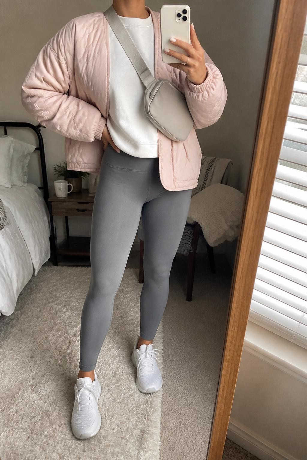 mirror selfie of a woman wearing the exact outfit described above, clearly showing a soft blush pink quilted jacket layered over an oversized white sweatshirt, gray high-waist leggings, white waterproof sneakers, a light gray belt bag worn crossbody, and silver stud earrings, holding a phone in front of a mirror, cropped at NECK so the face does not show, with a relaxed casual selfie pose, in a cozy bedroom mirror setup, cloudy morning light, with a coffee mug on a bedside table and a folded blanket on a chair, latest iPhone photo quality.