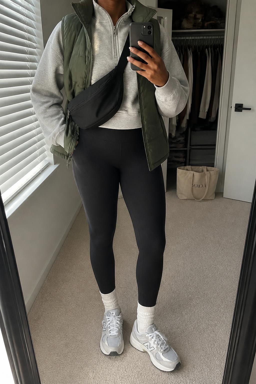Mirror selfie of a Black woman wearing matte black high-waisted leggings, an oversized heather-grey quarter-zip sweatshirt, an olive-green quilted puffer vest, cream crew socks, white and grey retro sneakers, and a black nylon crossbody bag, clearly showing the exact clothing items, colors, fabrics, and accessories listed, holding a phone in front of a walk-in closet mirror, cropped at the neck so the face does not show, relaxed casual selfie pose, natural window light, an open closet with clothes visible and a tote bag resting on the floor, casual and uncluttered, latest iPhone photo quality.