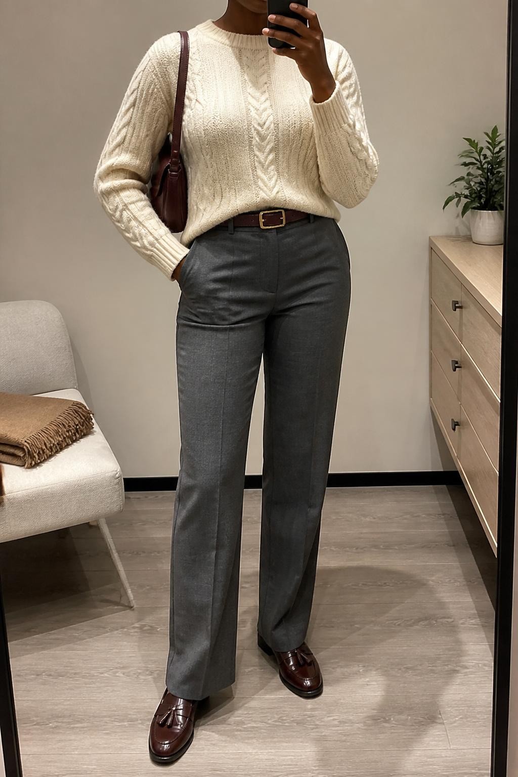 Mirror selfie of a Black woman wearing a cream cable-knit sweater, gray straight-leg work pants, burgundy loafers, a brown leather belt, and a burgundy shoulder bag, cropped at neck so the face does not show, in a minimal dressing room with soft indoor lighting, folded blanket on a chair and a small plant on a dresser, realistic latest iPhone photo quality.