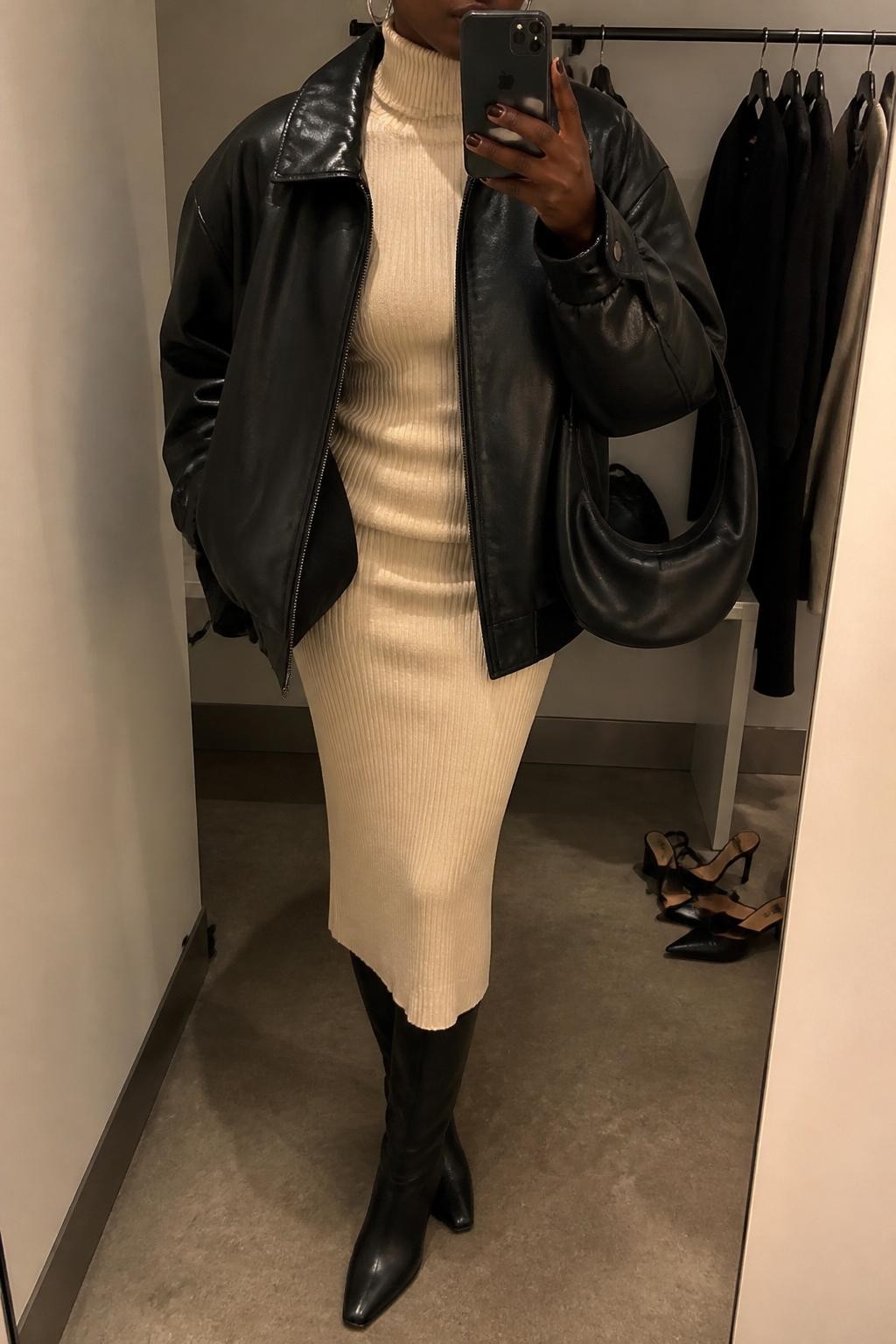 Mirror selfie of a Black woman wearing an oversized black leather jacket over a fitted cream ribbed turtleneck and matching cream ribbed midi skirt, clearly showing black knee-high leather boots, a soft black crescent shoulder bag, and chunky silver hoop earrings, holding an iPhone in front of a minimal dressing room mirror, cropped at the neck so the face does not show, relaxed casual selfie pose, warm moody lighting, a clothing rack with garments behind and a pair of heels on the floor nearby, natural unstaged setting, latest iPhone photo quality.