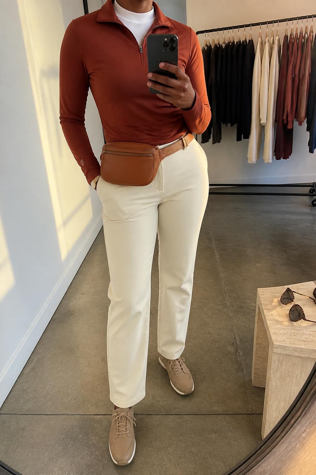 Mirror selfie of a Black woman wearing a rust fitted quarter-zip golf pullover over a white crew-neck performance top, paired with ecru straight-leg golf pants, clearly showing tan waterproof golf shoes, a cream cap, and a cognac belt bag worn around the waist, holding an iPhone in front of a fashion studio mirror corner, cropped at the neck so the face does not show, relaxed casual selfie pose, evening golden hour light, a clothing rack with garments behind and sunglasses resting on a side table, natural unstaged setting, latest iPhone photo quality.