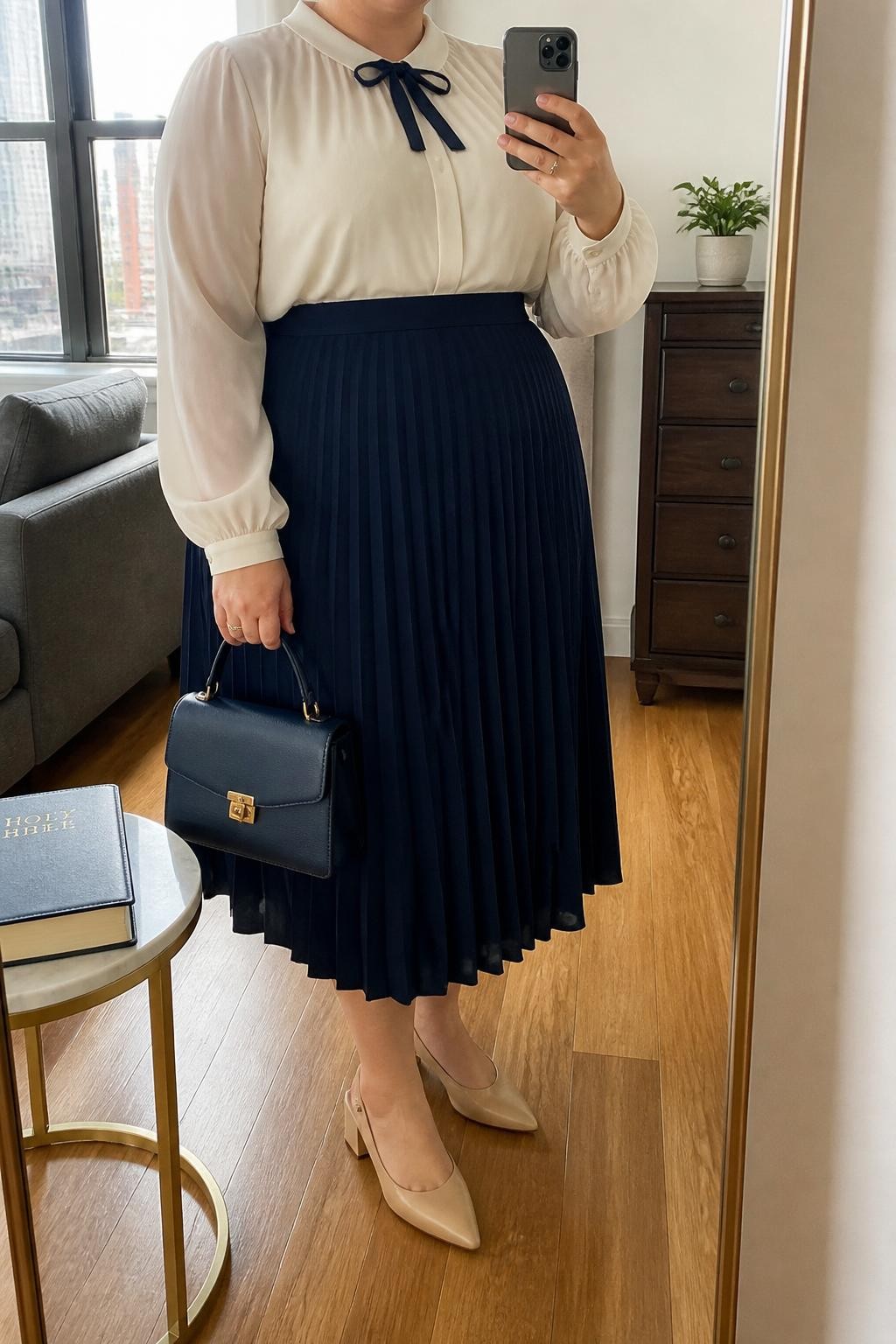 Mirror selfie of a plus size woman wearing a navy blue high-waisted pleated midi skirt, an ivory chiffon blouse with long sleeves and a small navy velvet bow at the neckline, nude pointed slingback heels with low block heels, a structured navy top-handle bag, and pearl stud earrings, clearly showing the exact clothing items, navy pleats, ivory chiffon, bow detail, footwear, and accessories listed, holding a phone in front of a mirror, cropped at the neck so the face does not show, relaxed polished selfie pose, in a stylish city apartment with natural window light, a Bible resting on a side table and a small plant on a dresser, natural unstaged church outfit, latest iPhone photo quality.