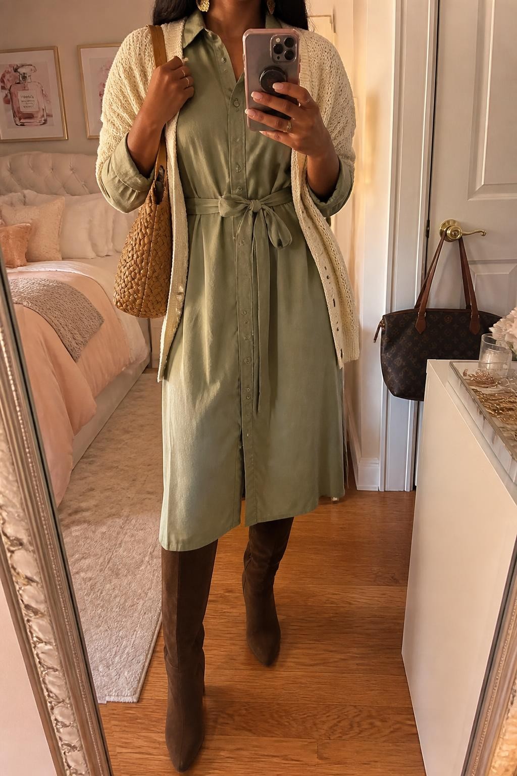 Mirror selfie of a Black woman wearing a sage-green cotton midi shirt dress with a matching waist tie, chocolate-brown suede knee-high boots, a cream knit cardigan draped over the shoulders, a woven tan shoulder bag, and hammered-gold earrings, clearly showing the exact clothing items, colors, fabrics, and accessories listed, holding a phone in front of a feminine-themed bedroom mirror, cropped at the neck so the face does not show, relaxed casual selfie pose, evening golden-hour lighting, a handbag hanging on a door handle and a jewelry tray on the vanity, warm and natural, latest iPhone photo quality.